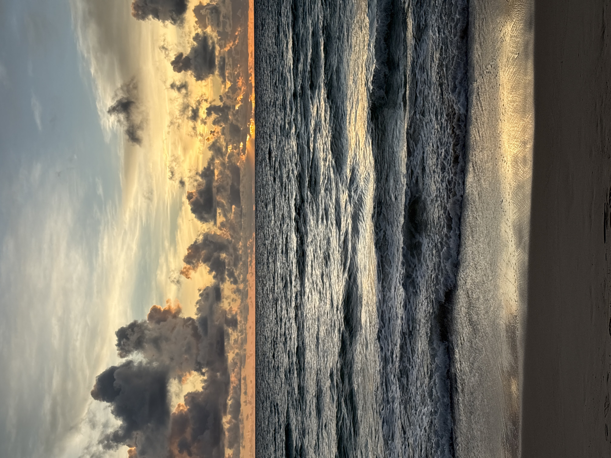 This media file depicts a serene beach scene at sunset in Choeng Thale, Thailand. The image is oriented vertically, dividing the frame into three distinct sections: sky, ocean, and sand.
The left side of the image features a dramatic sky filled with a mix of dark, brooding clouds and areas of bright, golden sunlight. The clouds are illuminated from behind by the setting sun, creating a warm glow with hints of orange and yellow against the fading blue sky. Wispy cirrus clouds are also visible, adding texture to the celestial canvas.
The central portion of the image is dominated by the ocean. Gentle waves are captured in motion, their crests white and foamy as they roll towards the shore. The water is a deep, dark blue-gray, reflecting the colors of the sky and the low-angled sunlight. The texture of the waves, with their individual ripples and troughs, is rendered in detail, suggesting a peaceful but active sea.
The rightmost section of the image showcases the sandy beach. The sand appears damp and smooth, reflecting the golden light of the sunset in a broad stripe. A dark shadow, possibly cast by an unseen object or dune, runs vertically down the right edge of the frame, contrasting with the illuminated sand.
The overall mood of the image is tranquil and evocative, capturing the beauty of a tropical sunset. The low light and dramatic cloud formations suggest it is late evening. There is no text visible in the media file.