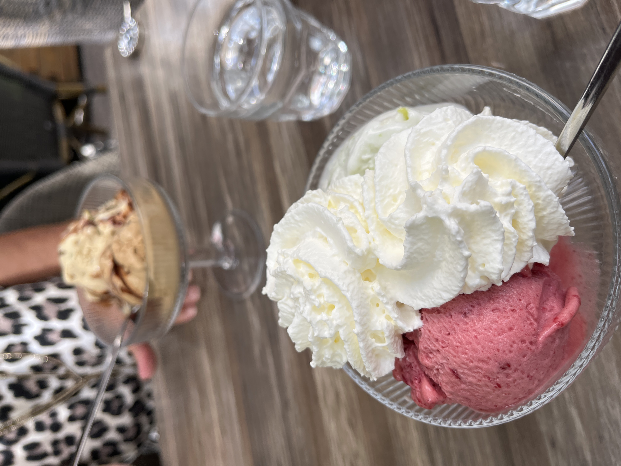 A close-up shot focuses on a clear glass bowl filled with two scoops of ice cream, topped with a generous swirl of whipped cream. One scoop is a vibrant pink, suggesting a berry flavor, while the other, mostly obscured by the whipped cream, appears to be a lighter color. A person's hand, adorned with a leopard-print garment and a delicate necklace, is visible in the upper left, holding a clear plastic spoon. The spoon is poised above a second clear glass bowl in the background, which contains a scoop of light brown ice cream, possibly coffee or caramel flavored, with visible caramel drizzle.
The scene is set on a wooden table. To the right of the main bowl, a clear glass of water is visible, with light glinting off the water's surface. In the background, out of focus, a person is seated at another table, and the overall ambiance suggests an indoor dining setting, possibly a cafe or restaurant. The lighting is natural and diffused, highlighting the textures of the ice cream and whipped cream. No specific landmarks or indicators of Montélimar, France, are present in the image.