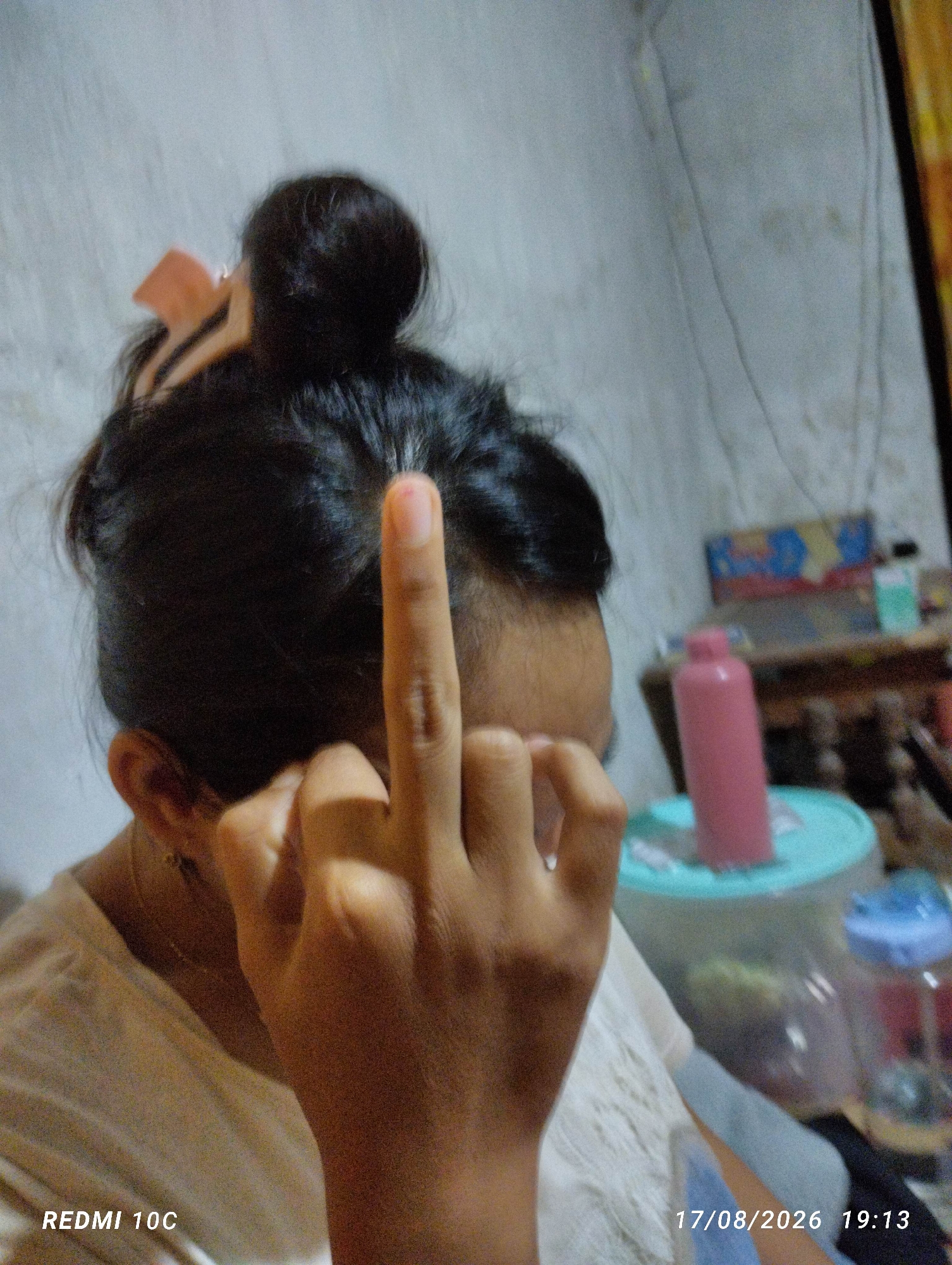 A person's left hand is raised, with the middle finger extended, in the foreground. The person is wearing a light-colored t-shirt. Their hair is pulled up in a bun. The background is blurred and shows a room with furniture, including a pink bottle and containers. The timestamp "17/08/2026 19:13" is visible in the bottom right corner. The text "REDMI 10C" is visible in the bottom left corner. The image was taken in Kembiritan, Indonesia.