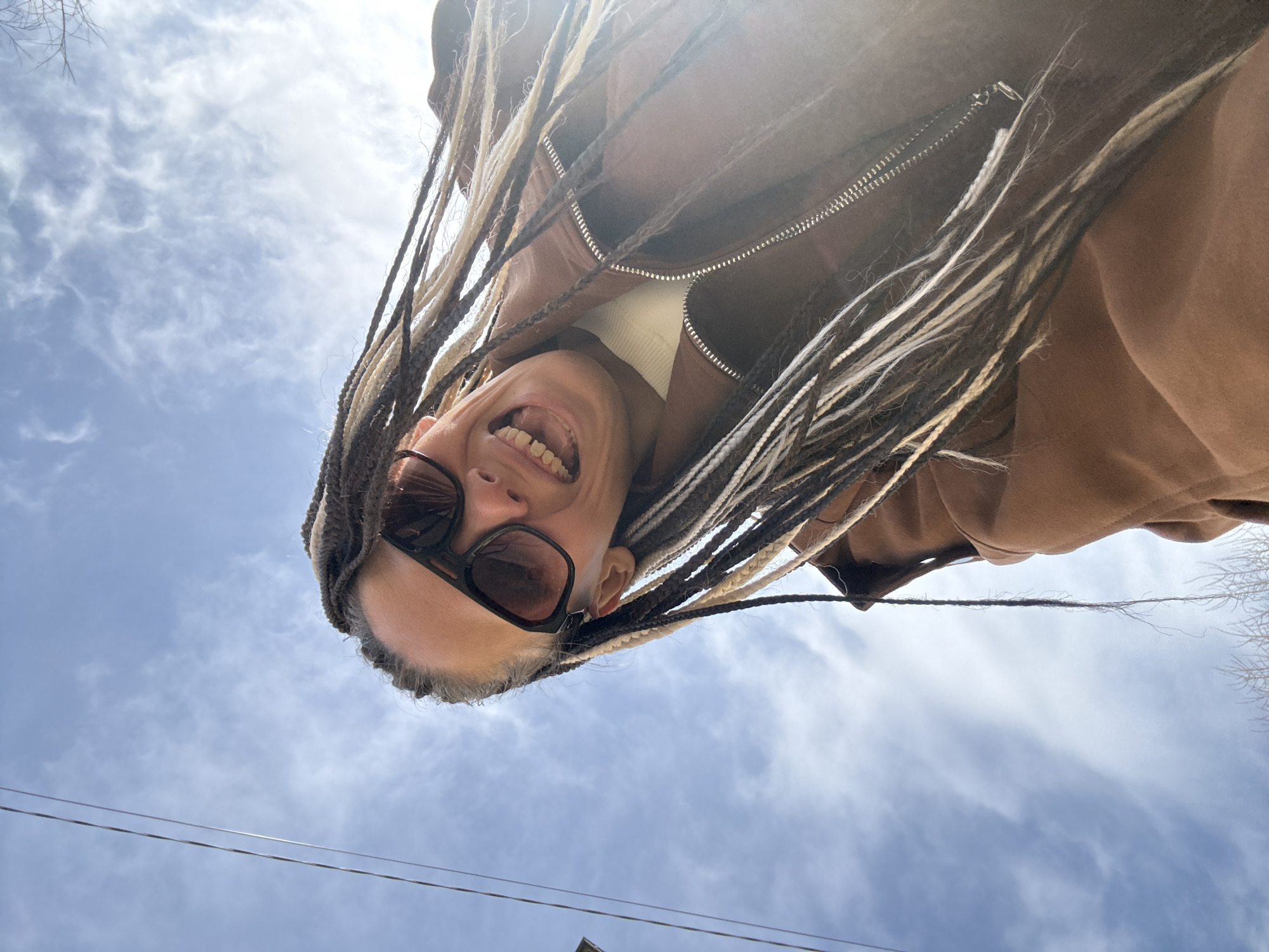 The image is taken from a low-angle perspective, looking up at a person with light brown and white braids, wearing sunglasses, a white turtleneck, and a brown jacket. The person is smiling widely, exposing their teeth. The background shows a partly cloudy blue sky. In the lower part of the frame, there are some power lines and the top of a building. There's no definitive evidence to confirm the location as Rusanovo, Russia, based solely on the visual information provided in the image.