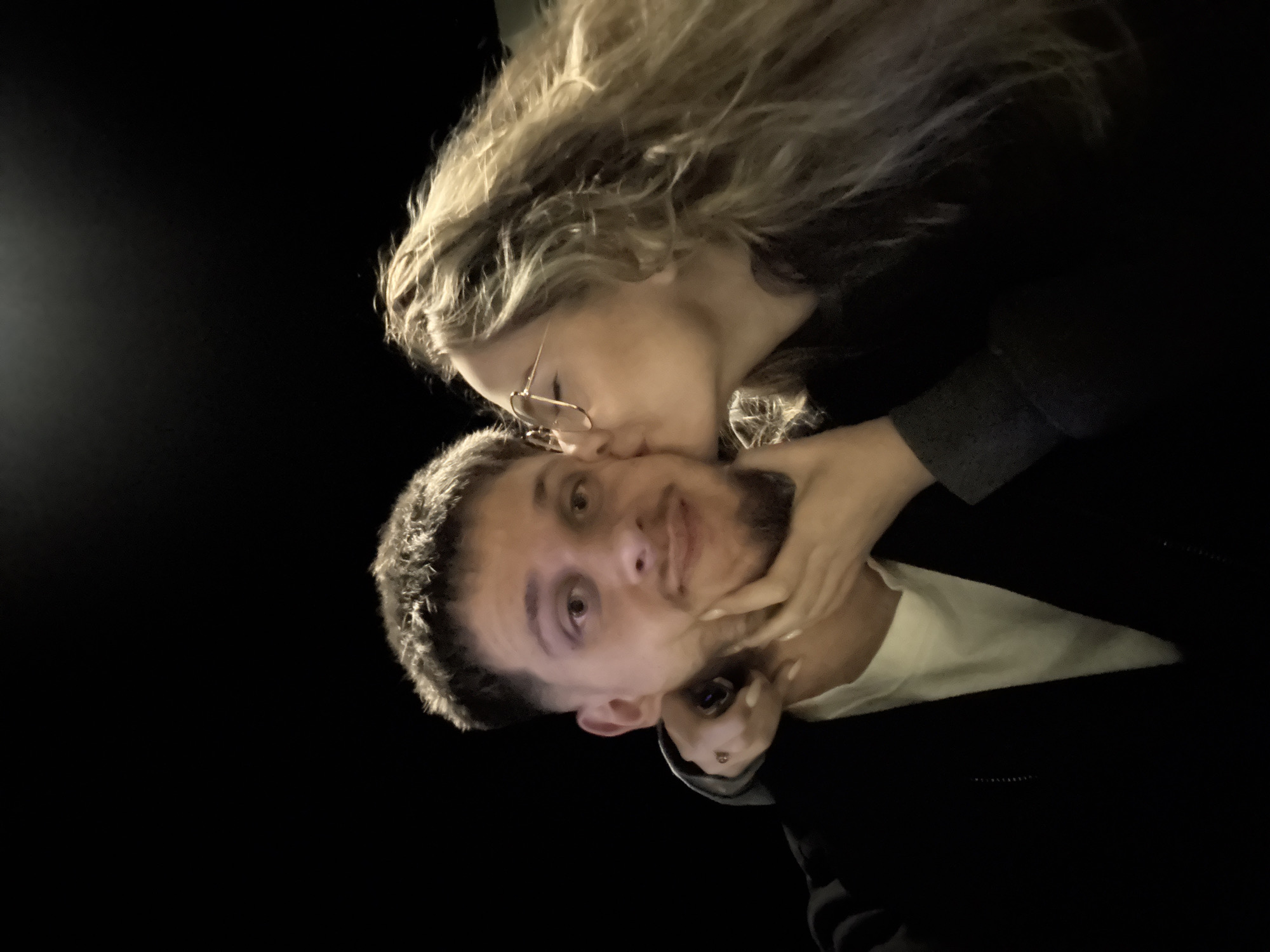 A woman with wavy, blonde hair and wearing glasses is kissing a man on the cheek. The man has short, dark hair and a beard. They are both wearing dark clothing. The photo appears to be taken at night, with a dark background. The location of Blagoevgrad, Bulgaria, cannot be determined from the visual information provided.