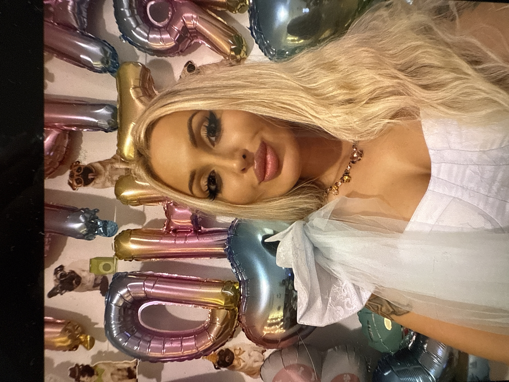 A blonde woman is captured in a close-up, eye-level portrait, posing in front of a decorative wall. She has long, wavy hair, dark eye makeup, and pink lipstick, looking directly at the camera. She wears a light-colored, possibly white, top or dress with a textured or ruffled sleeve and a multi-stone necklace with amber-toned beads. A tattoo is faintly visible on her right arm.
The background features a light-colored wall adorned with numerous metallic foil balloons in iridescent, gold, pink, and blue shades. These balloons spell out the word "BIRTHDAY" vertically, with discernible letters including "B", "I", "R", "T", "H", "D", "A", and "Y". Other balloons that appear to be numbers, possibly "1" and "8" or "8" and "1", are also part of the arrangement, suggesting a birthday celebration. Among the balloons, small decorative elements resembling cartoon animals, including a pug dog, are attached to the wall. A small, square, green object, possibly an old music device, is also present on the wall. The setting appears to be an indoor space, brightly lit for the occasion.