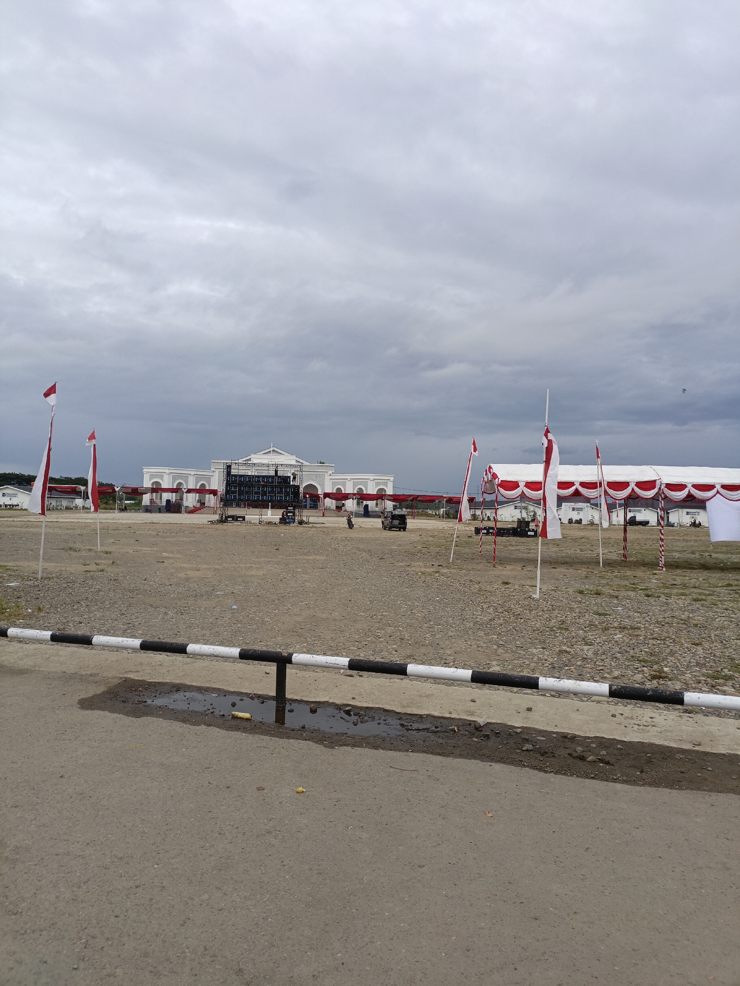 The image depicts an outdoor scene with a cloudy sky. In the background, there is a large white building with arched windows and decorative white columns. In front of the building, a temporary stage structure with numerous speakers is set up. To the right of the stage, there are several white tents with red trim and poles. Various Indonesian flags, red and white, are planted in the ground throughout the area. The ground is covered with gravel and some asphalt in the foreground. A black and white striped barrier runs across the bottom of the image, with a puddle of water near it. There are a few vehicles and people visible in the distance, including someone on a motorcycle and individuals on the stage structure. The scene appears to be set up for an event or festival. The location is identified as Meureudu, Indonesia, suggesting it is likely a public space or plaza.