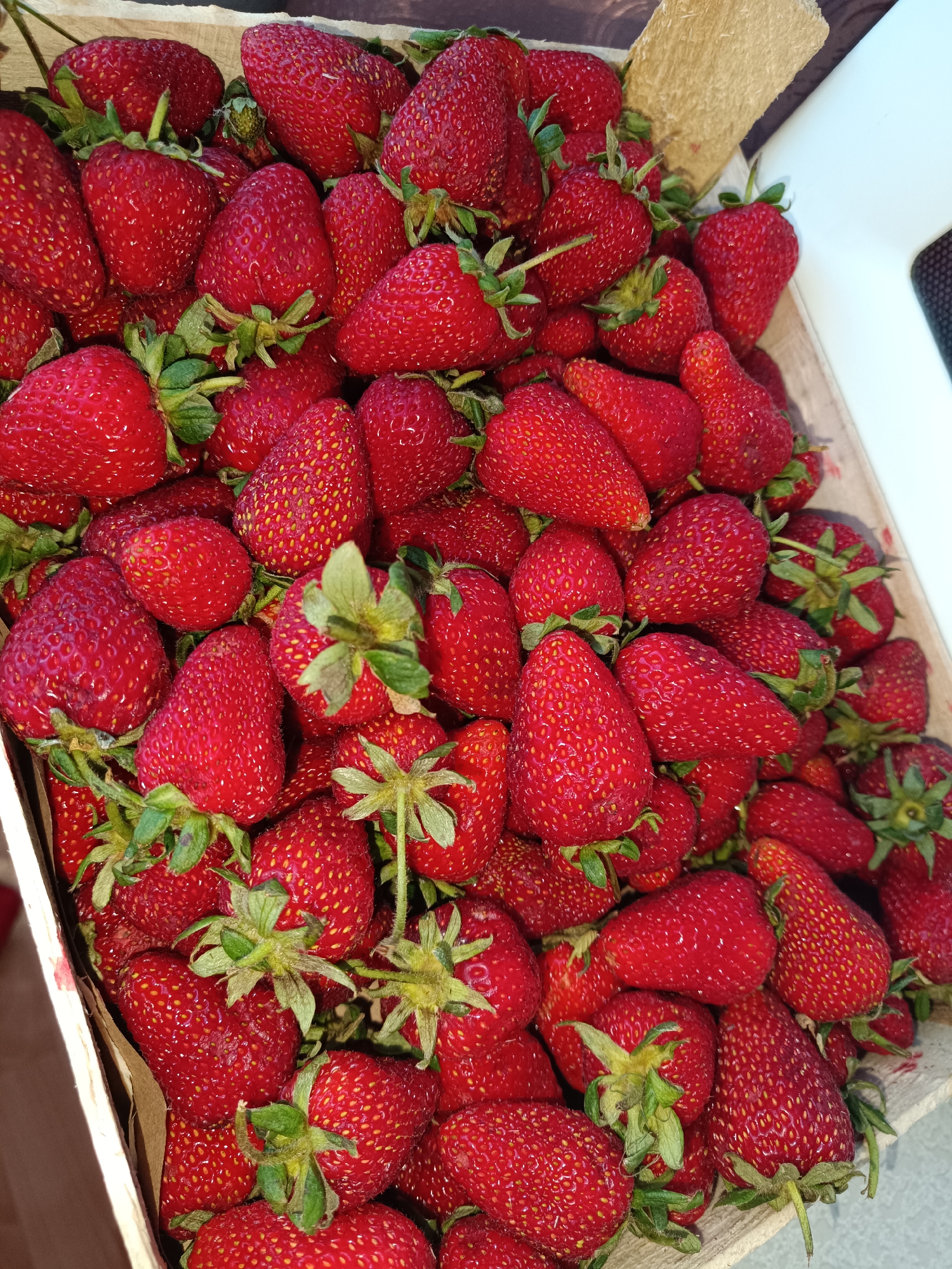 A high-angle, close-up shot depicts a wooden box brimming with fresh, ripe strawberries. The strawberries are predominantly red and of varying sizes, with green stems visible among them. The box appears to be made of light-colored cardboard. The location is not discernible from the image; therefore, the location in Tobolsk, Russia, cannot be confirmed based on visual evidence. No people or actions are visible. The environment consists solely of the box and the strawberries.