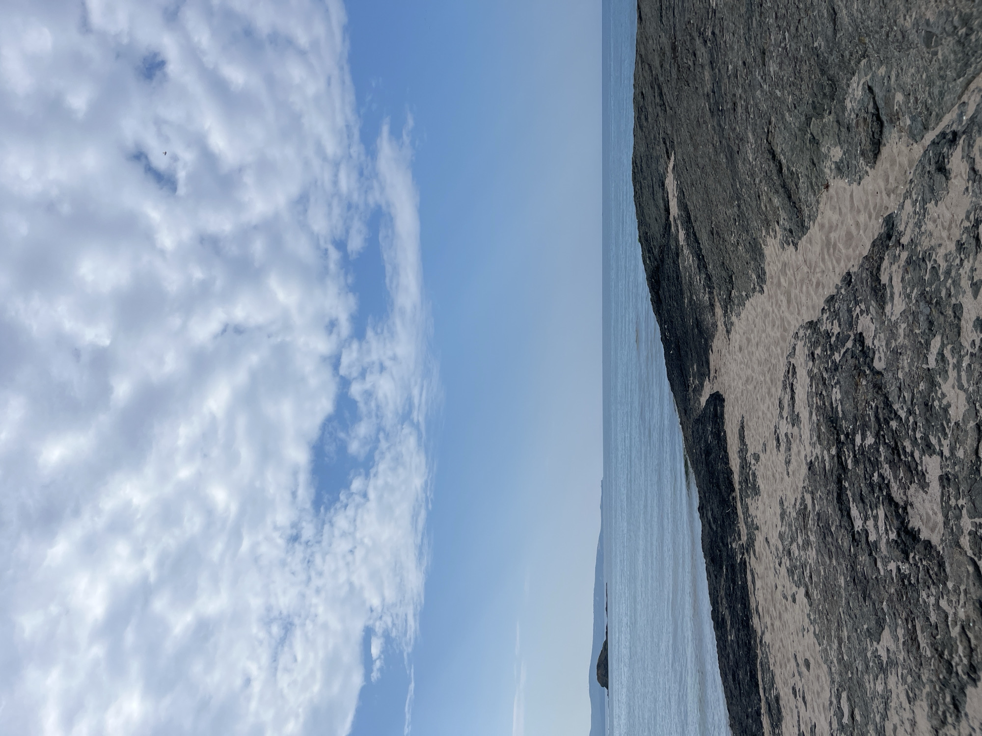The image depicts a coastal scene at Lozenets, Bulgaria. In the foreground, a rocky shoreline with patches of sand leads to the sea. The water is calm and a light blue, meeting the horizon under a sky filled with scattered white clouds against a clear blue background. In the distance, landmasses are visible on the horizon. The overall scene is natural and serene, with no visible human activity.
