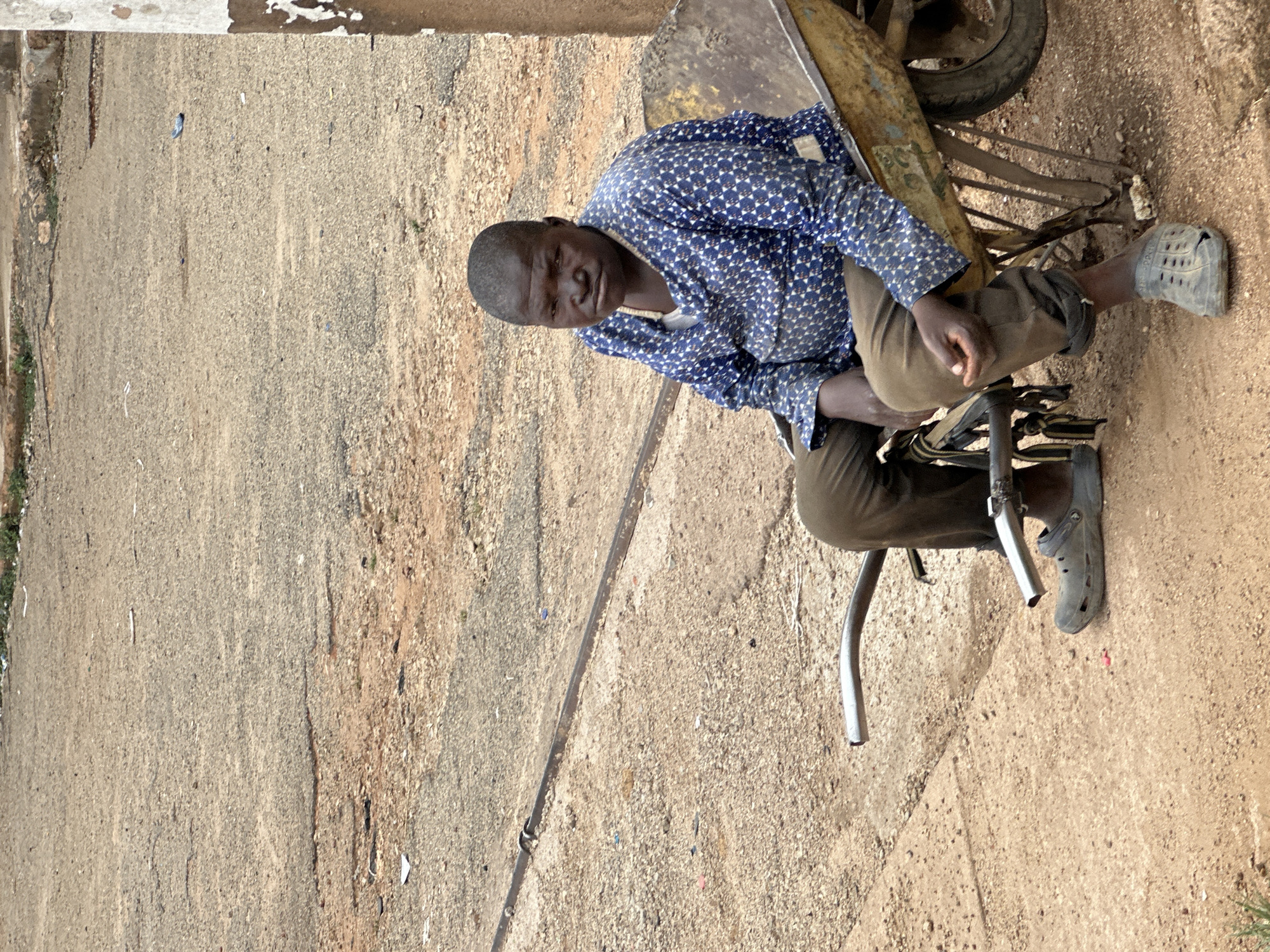 An adult male is captured in an outdoor, eye-level shot in Gura Top, Nigeria. He has dark skin, short dark hair, and is wearing a long-sleeved blue shirt with a white geometric pattern, dark trousers, and grey perforated clogs. He is seated on a low, dark metal frame or stool, with his body angled slightly towards the right and his face turned to look directly at the camera with a calm, neutral expression.
Behind and to the right of the man, part of an old, yellow or gold wheelbarrow with a single visible wheel is propped up. The ground beneath him and extending throughout the frame is a dry, textured mix of dirt, sand, and small gravel, typical of an unpaved road or pathway. Scattered across the ground are various small pieces of debris and litter. To the left, a linear demarcation suggests a subtle change in ground texture or a shallow drain. The scene is brightly lit, indicating clear, sunny daylight. There is no visible text in the image.