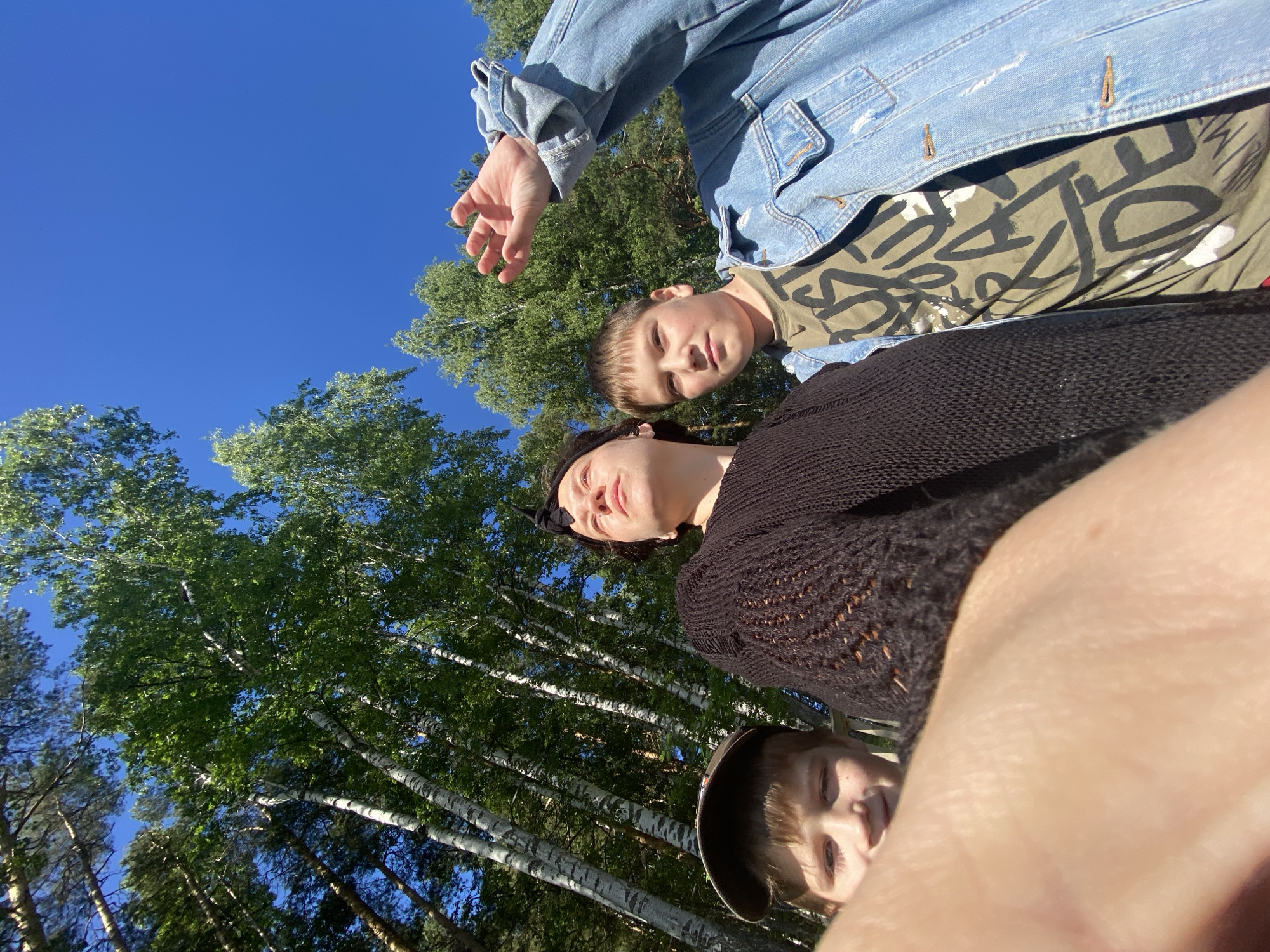 In a sunlit outdoor setting, possibly a park or wooded area in Chaikovsky, Russia, three individuals are captured in a low-angle selfie. The sky above is a clear, vibrant blue, and tall, slender birch trees with green foliage dominate the background, suggesting a summer day.
In the foreground, partially obscuring the scene, is a close-up of a hand or arm, appearing to hold the camera. To the left, a young boy with light brown hair, wearing a brown baseball cap, is visible from the chest up. He has fair skin and is looking slightly to his left, with one eye squinted.
To the right of the boy and slightly behind him is a person with dark hair, wearing a black, loosely knit sweater. This individual is looking upwards and to the left with a contemplative expression.
Further to the right and behind the person in the black sweater is another young individual, appearing to be a boy, wearing a light olive-green t-shirt with bold black lettering and a light blue denim jacket. He is looking directly at the camera with a neutral expression and has his right arm raised, with his hand open, reaching towards the top of the frame. The overall atmosphere is casual and bright.