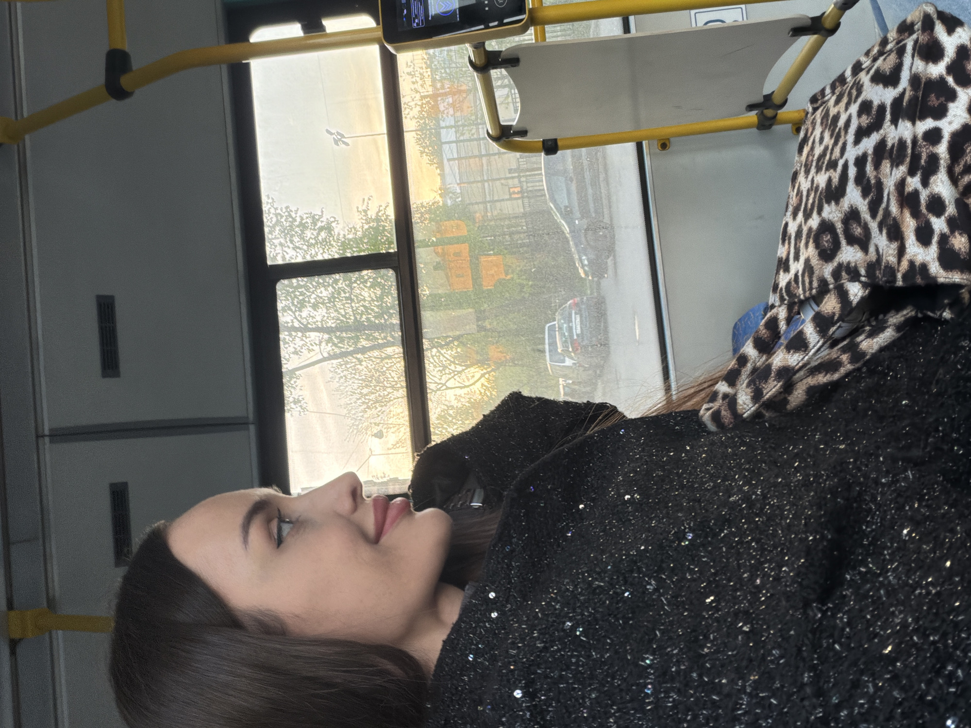 A woman is seated on a bus, looking out the window at Saint Petersburg, Russia. The exterior view shows buildings, trees without leaves, streetlights, and a row of parked cars. The bus interior includes a yellow handrail, a window shade, and another passenger's leopard print jacket. The woman has brown hair and wears a dark, possibly sequined, top.