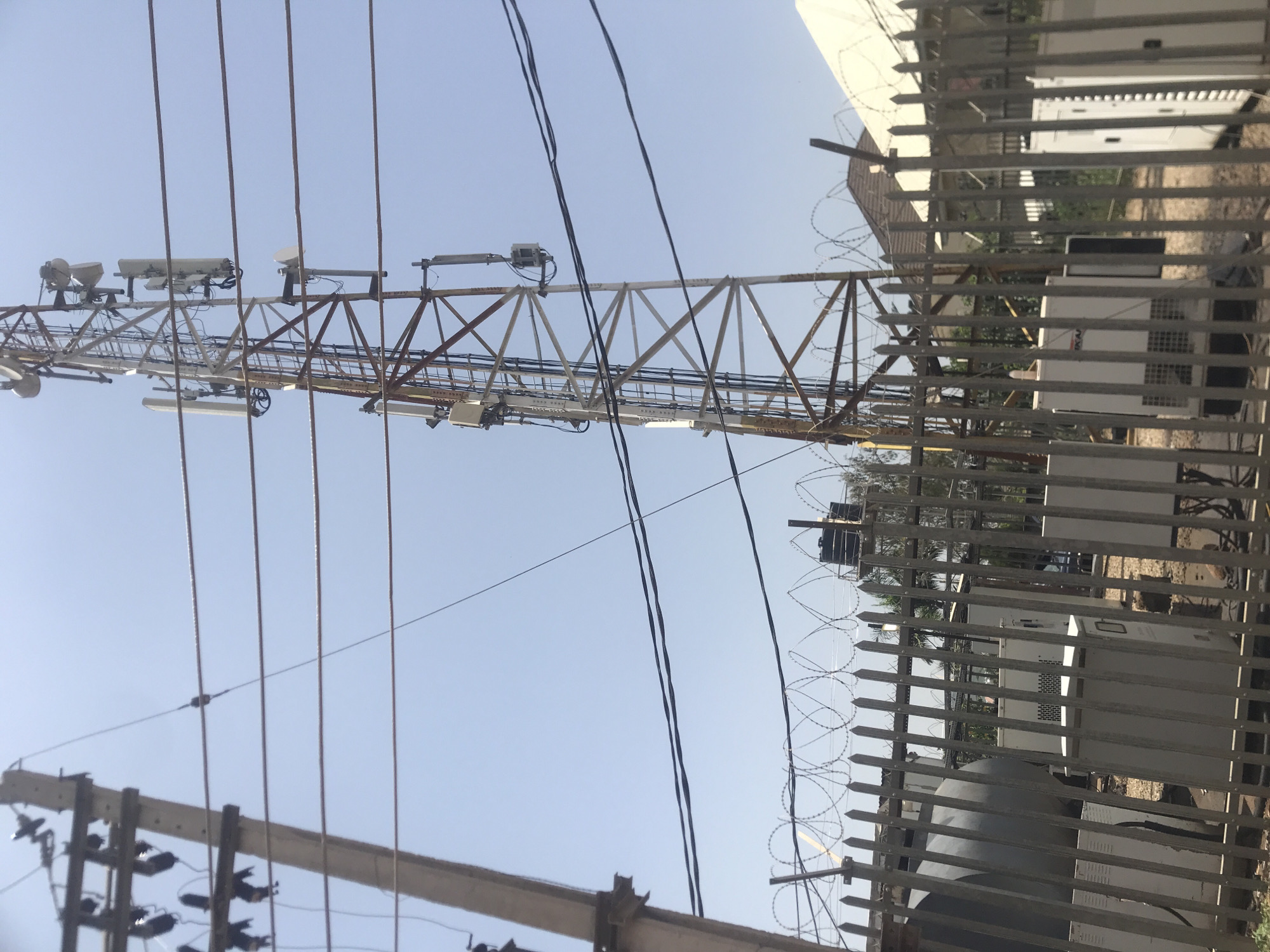 The image depicts a telecommunications tower in the city of Rafin Par, Nigeria. The tower is a lattice structure with multiple antennas mounted at various levels, suggesting it serves as a cell tower. Below the tower, a metal fence with barbed wire runs across the foreground, separating the viewer from a compound. Within the compound, several pieces of equipment are visible, including large grey cylindrical tanks and white rectangular enclosures that appear to be electrical or cooling units, possibly associated with the telecommunications equipment. A concrete utility pole with multiple power lines is also present on the left side of the image, adding to the industrial and technological infrastructure evident in the scene. The sky is clear and blue, indicating a daytime setting. The overall environment appears to be an urban or semi-urban area in Nigeria, characterized by functional infrastructure and a clear, bright sky.