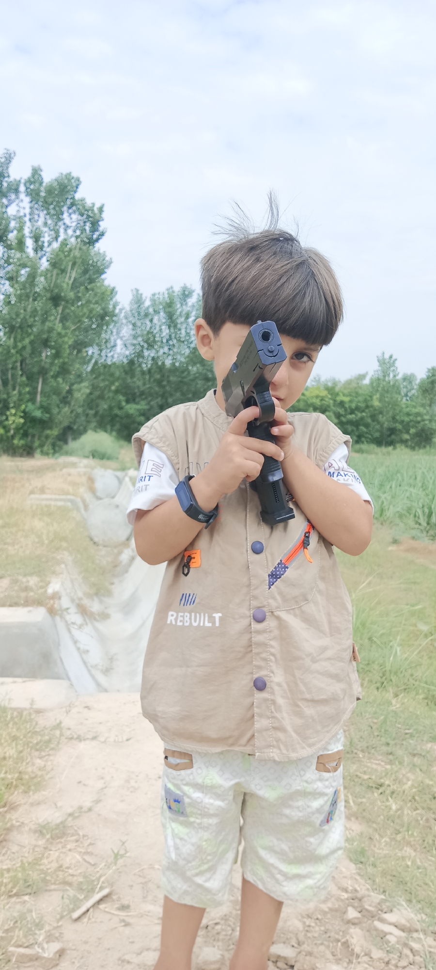 A young boy is standing outdoors, holding a black toy gun in his hands, aiming it forward. He is wearing a light brown vest over a white t-shirt, and white shorts with a patterned print. His hair is brown and he has dark eyes. The background consists of green trees and a cloudy sky. A dirt path and some concrete structures are visible on the left side of the image. The scene appears to be in a rural or semi-rural area, possibly on the outskirts of Katlang Tehsil, Pakistan.