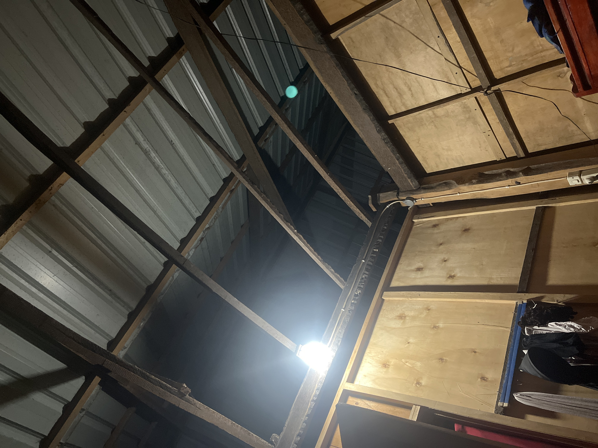 The image depicts the interior of a structure with a corrugated metal roof supported by wooden beams. Some sections of the roof appear damaged or incomplete, allowing light to enter. A bright white light bulb hangs from the ceiling, illuminating the area. The walls are constructed of large wooden panels. On the left wall, a rack with several items of clothing is visible. On the right wall, electrical wiring and a wall socket are present. In the lower right corner, a portion of a red wooden object, possibly a piece of furniture, is visible. The scene appears to be an indoor space in Turumame, Indonesia, possibly a workshop or storage area.
