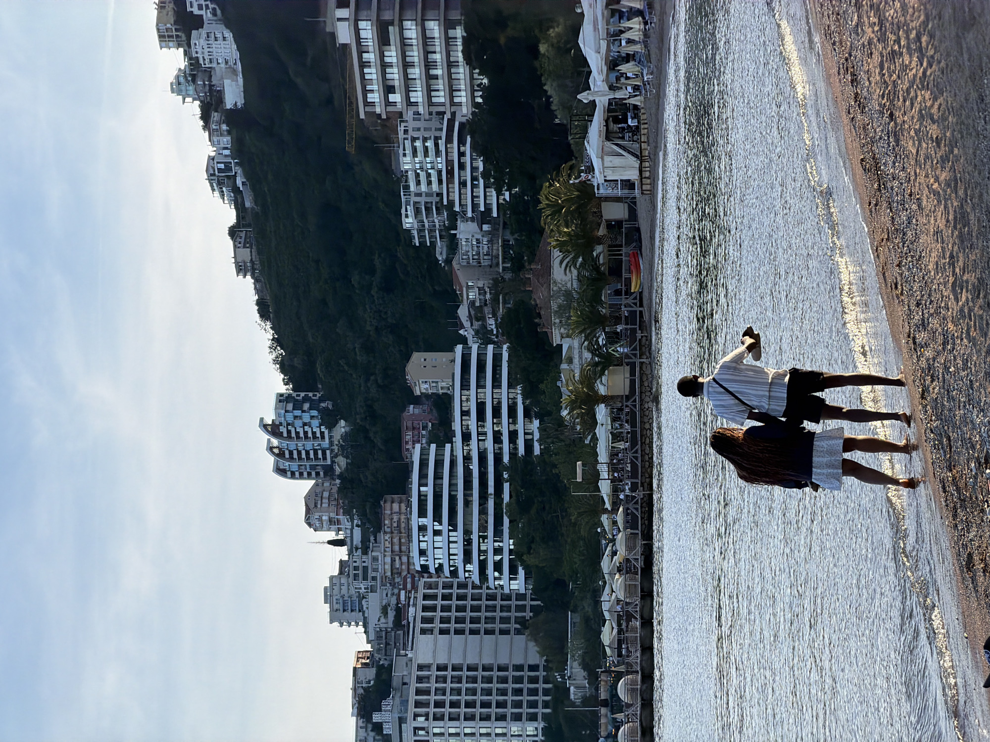 A beach scene in Budva, Montenegro, shows a couple walking along the shore, with their backs to the viewer. The man wears a striped shirt and dark shorts, while the woman has long hair, a dark jacket, and a light-colored skirt. Behind them are buildings on a hillside, and in the distance, more buildings are scattered along the coast. The water is calm with small waves, and the beach is sandy with a visible shoreline.
