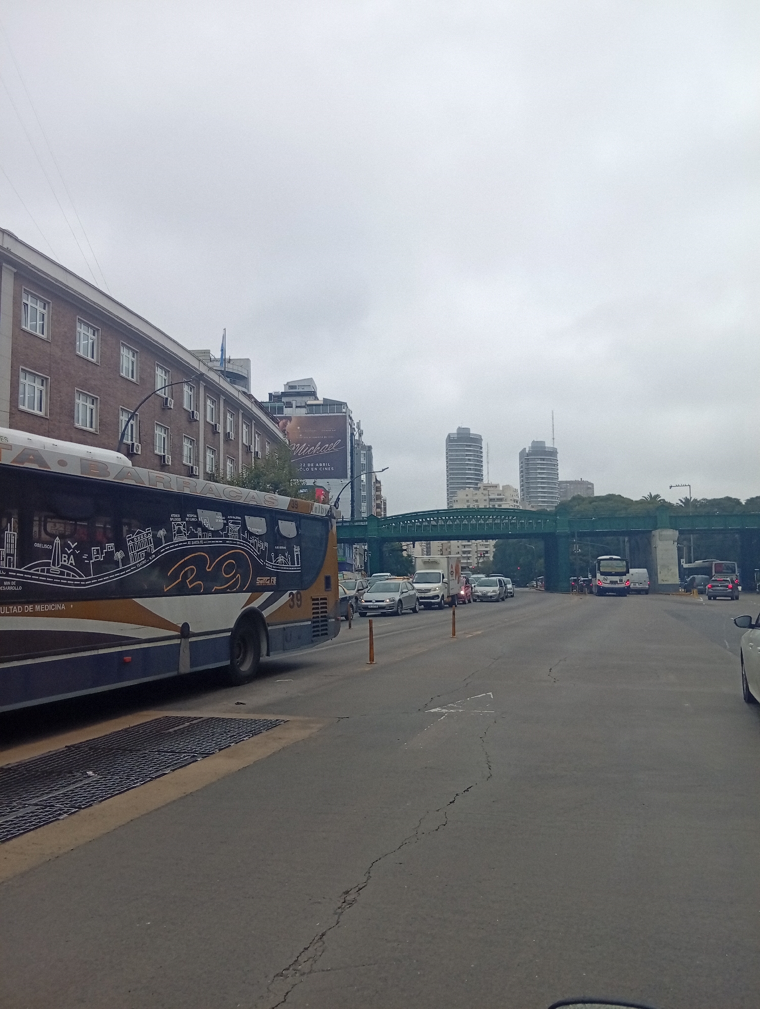 The image depicts an overcast urban street scene in Buenos Aires, Argentina, characterized by vehicular traffic and diverse architecture under a grey sky. In the foreground on the left, a large blue and yellow city bus, identified as Route 39 and bearing the destination "BARRACAS," is prominently featured. Its side showcases a graphic with "OBELISCO," "BA," and "FACULTAD DE MEDICINA," along with other points like "HOSPITAL" and "ALTO PALERMO," confirming the city.
The road is filled with various cars and vans, suggesting ongoing traffic or a busy commute. In the middle ground, a distinctive green elevated railway or bridge structure spans across the street. Behind it, modern high-rise buildings rise into the cloudy sky. To the left, a traditional multi-story brick building with many windows lines the street. Above some buildings, a billboard advertises "Michael" playing "22 DE ABRIL SÓLO EN CINES." The street surface shows some cracking. The overall impression is that of a typical cloudy day in a bustling South American city.