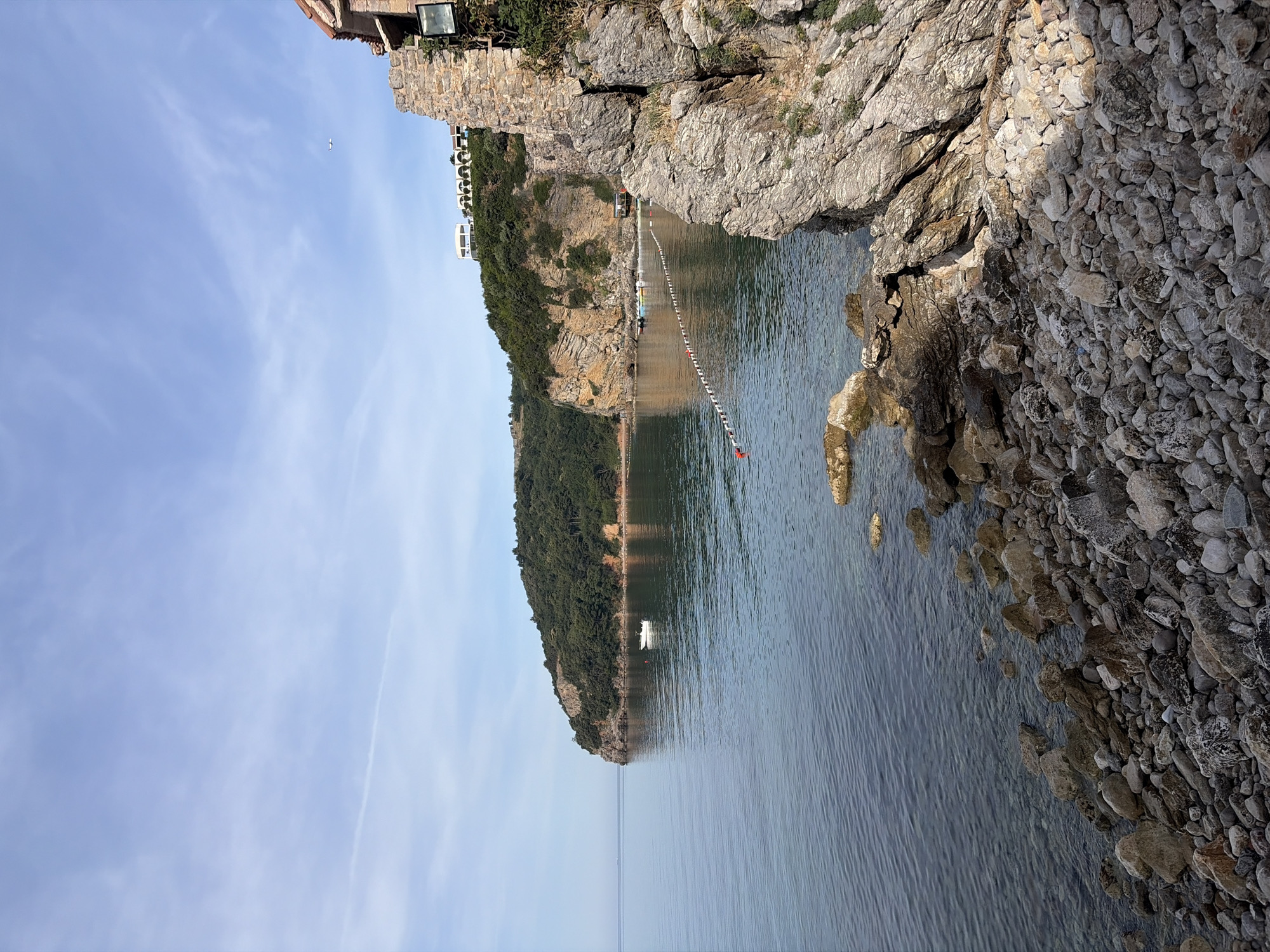 Captured from a high vantage point, the image depicts a cove in the city of Budva, Montenegro, surrounded by rocky cliffs and lush greenery.
On the right of the frame, a rocky shoreline is visible, comprising gray and beige stones and boulders. To the left, a cliff face ascends, showcasing a mix of exposed rock and dense green vegetation. At the cliff's edge, there is a cluster of structures, possibly residential or tourist accommodations.
The cove itself is filled with water that ranges in color from deep blue to a lighter turquoise as it approaches the shore. A string of buoys stretches across the water, marking a boundary, perhaps for swimming or boating.
In the distance, a peninsula covered in vegetation extends into the water, creating a sense of depth. A single white boat sits near the peninsula. The sky above is a light blue hue, with scattered clouds adding texture to the scene.
There are no people visible in the image.