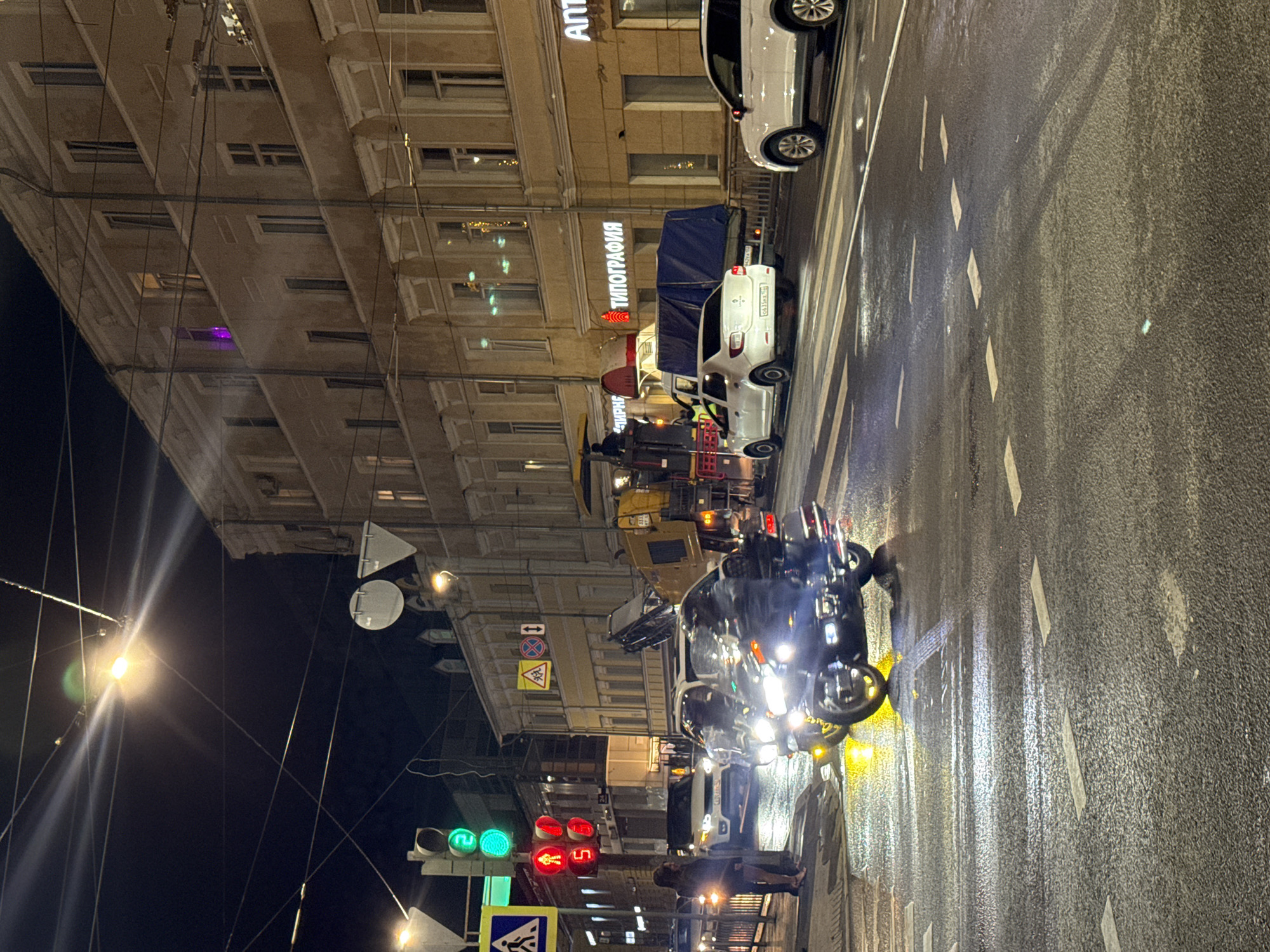 Captured at night, the image depicts a street scene in Nizhny Novgorod, Russia. The street is wet, reflecting the city lights and vehicle headlights. Multiple vehicles are visible, including cars and what appears to be a utility truck. Pedestrians are present on the sidewalk. The buildings lining the street exhibit architectural details. A sign reads "ТИПОГРАФИЯ," and another contains the Cyrillic alphabet "АПТ". Traffic lights are visible, showing green and red. Street signs, including crosswalk signs, are also in view. The image is shot at an angle.