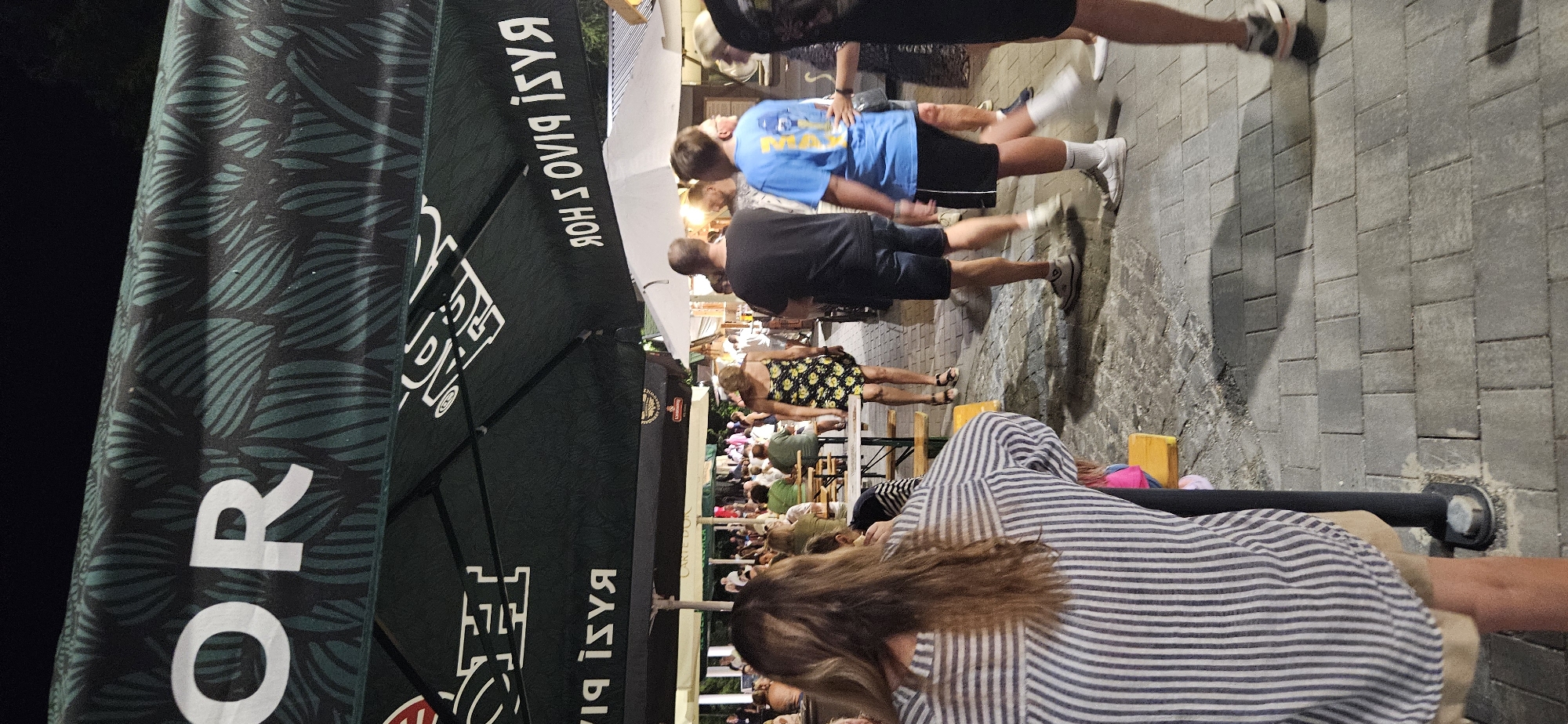 The image depicts an outdoor scene at dusk, likely in Hévíz, Hungary, judging by the Hungarian text on the umbrella. A large, dark green umbrella with a leaf pattern and white lettering is prominently featured, casting shadows on the paved ground. The umbrella appears to be associated with a food or beverage establishment, as indicated by visible logos and text like "PIVO" (beer in Slavic languages) and "HOR".
In the foreground, a person with long, light brown hair is seated at a table, wearing a striped shirt and shorts. Their back is to the camera, and they are partially obscured by a pole.
Further back, several other individuals are visible. A woman in a yellow floral dress walks away from the camera. Two men are also seen walking, one in a black t-shirt and denim shorts, and another in a blue t-shirt and black shorts. Other people are seated at tables in the background, suggesting a social gathering or dining area. The environment appears to be a pedestrian walkway or outdoor seating area with cobblestone paving. The lighting suggests it is evening, with artificial light sources illuminating the area.