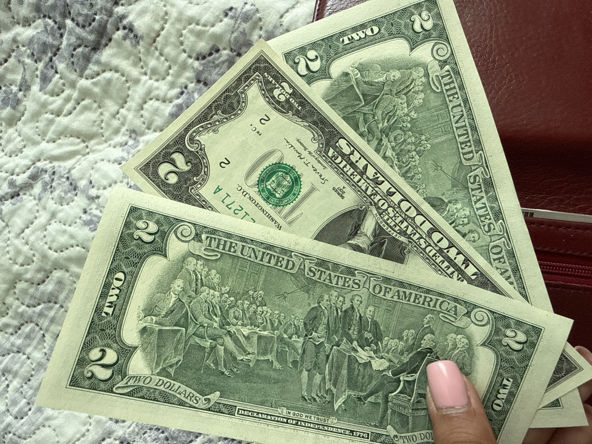 The image shows a close-up of several US two-dollar bills fanned out on a textured, light-colored surface. A person's hand with a pink-painted fingernail is visible on the left side, holding the bills. The bills are green and feature engravings of historical scenes and text. The background surface appears to be a quilted fabric with a subtle floral pattern. There is no indication of the location being Ekzarh Antimovo, Bulgaria, or any urban environment. The scene is static and appears to be a simple display of currency.