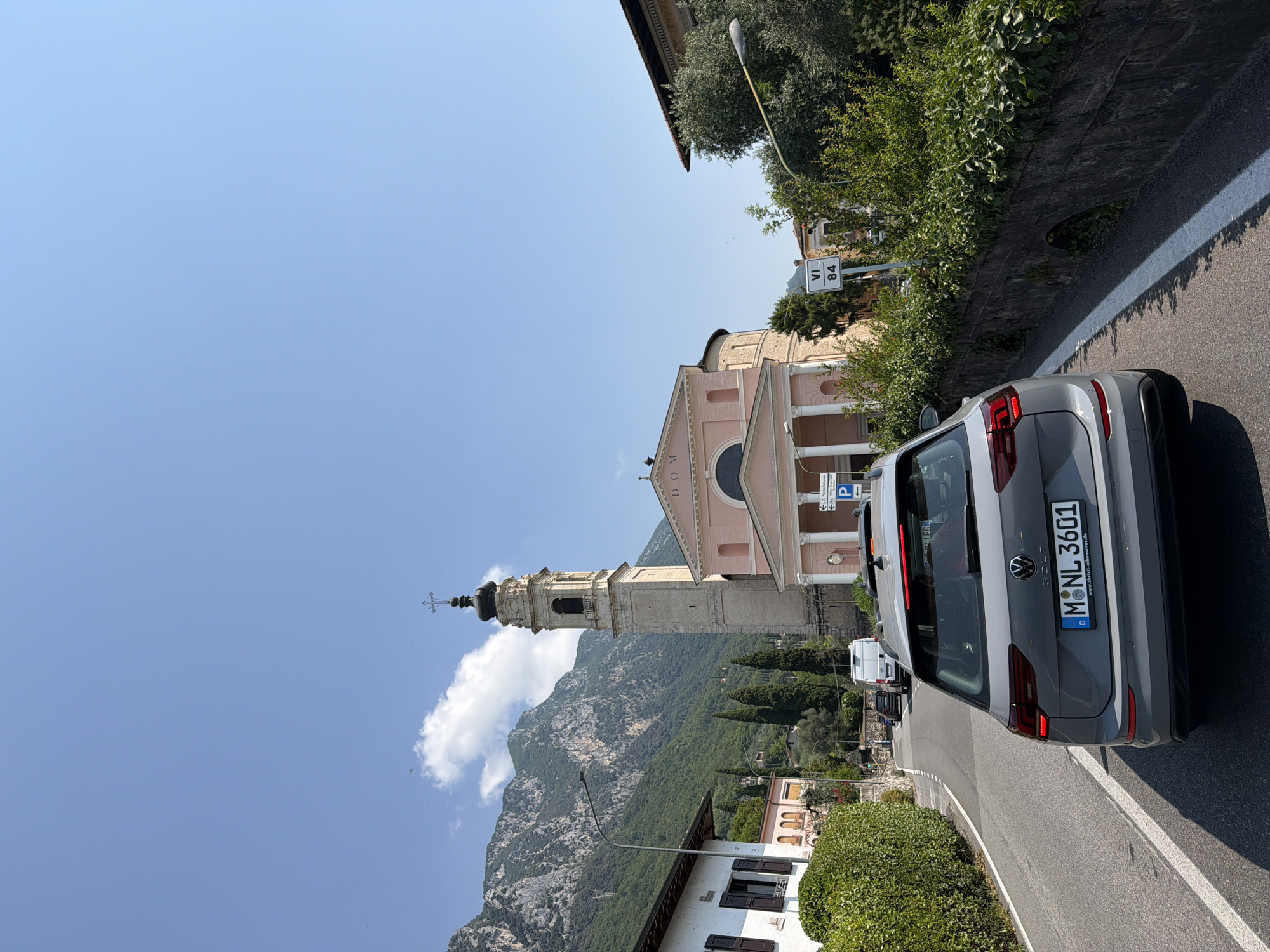 The image shows a gray Volkswagen Golf driving on a paved road in Gargnano, Italy. The car is in the foreground, facing away from the camera. In the background, there is a pink church with a tall bell tower. Mountains and lush green trees surround the church. The sky is clear and blue with a few white clouds. There are other cars and buildings visible on the road, as well as a street lamp. A road sign reads "VI 84".