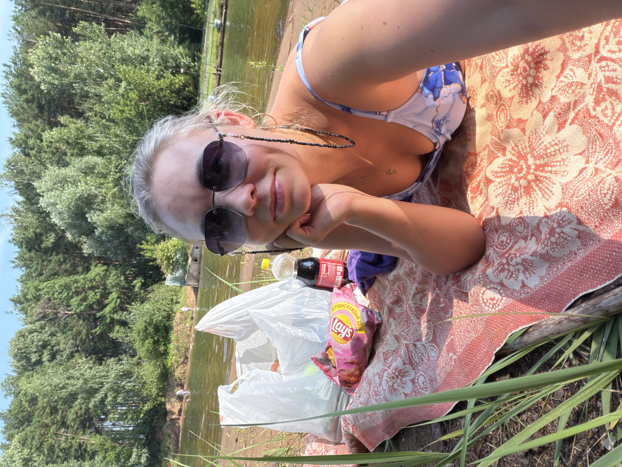 The image captures a woman reclining on a blanket by a body of water, likely a reservoir or river, in the city of Chaikovsky, Russia. She is wearing sunglasses, a blue and white patterned swimsuit, and a necklace. Next to her, on the blanket, are two white plastic bags, a bag of Lay's chips, a bottle of dark soda, and a folded purple towel. The background features a dense forest bordering the water, with a clear blue sky above. Several people can be seen in the water or on the shore in the distance, suggesting a public recreational area. The overall scene depicts a relaxed outdoor activity, possibly a picnic or day at the beach.