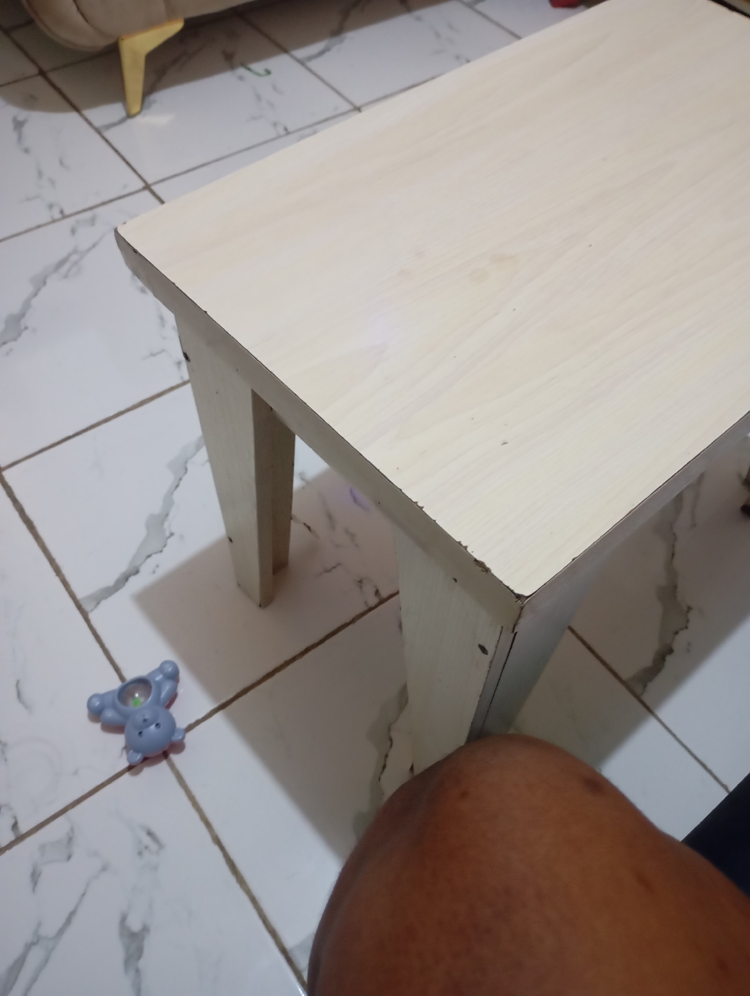 An overhead shot captures a light-colored wooden table situated on a tiled floor. The table has a simple, rectangular design with four legs. To the left of the table, on the floor, lies a small, light blue toy. The floor is composed of large, marble-patterned tiles. In the bottom right corner of the frame, a portion of a person's leg is visible, suggesting the presence of an individual in the scene. The lighting appears to be ambient, casting subtle shadows.