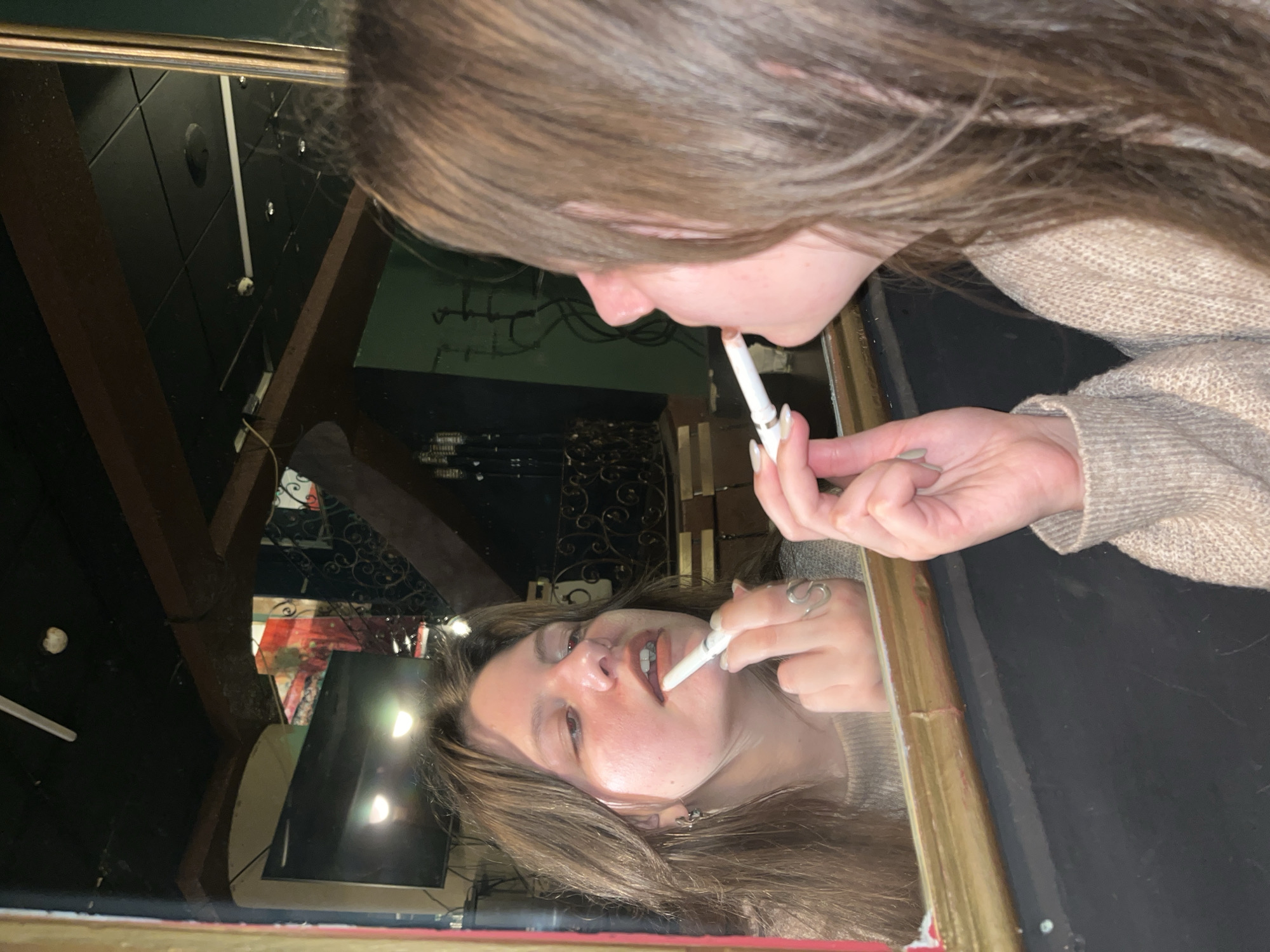 A young individual, appearing female, with medium-brown hair and a light beige knitted sweater, is depicted in a close-up, reflected in a rectangular mirror with a worn gold-toned frame. The subject is applying a brownish-pink cosmetic to their upper lip with a white cylindrical applicator. A silver-toned, wavy-shaped ring is visible on the middle finger of the reflected left hand, and small earrings are present on the reflected left earlobe. The fingernails are painted a light, neutral color. The reflection reveals the interior of what appears to be a public establishment, such as a bar or restaurant, located in Varna, Bulgaria. The background features a dark ceiling with square tiles, a fluorescent light fixture, and a small, round white object consistent with a smoke detector or security camera. Prominent dark wooden beams crisscross the ceiling. A wall-mounted television screen, dark green wall sections, decorative wrought iron work, and a colorful tapestry or wall hanging with red and green hues are visible in the background reflection, suggesting a dimly lit, rustic interior.