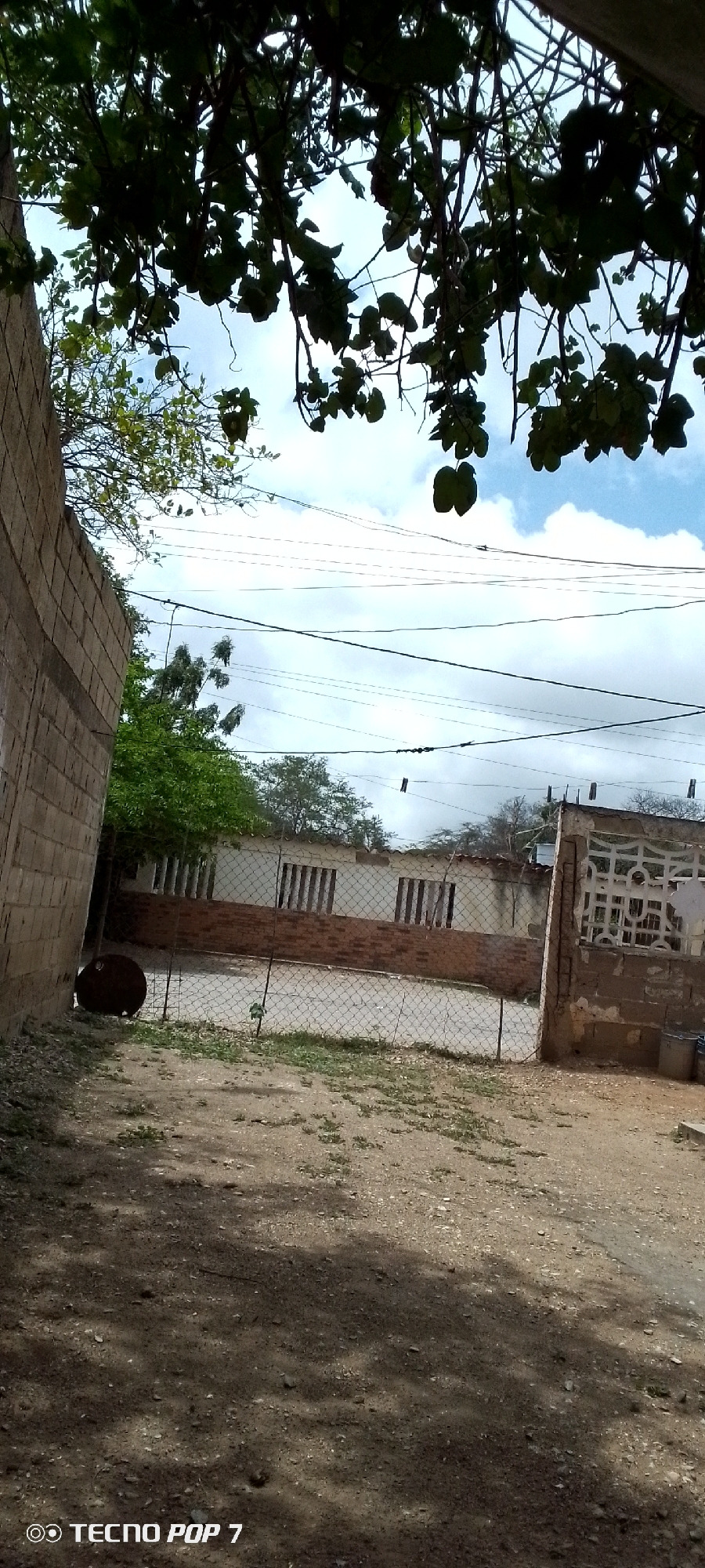 The image depicts an outdoor scene in what appears to be a rural or semi-urban setting. In the foreground, a dirt path with scattered pebbles and some sparse vegetation is visible, shaded by the leaves and branches of trees overhead. To the left, a wall constructed from concrete blocks lines the edge of the path. A large, dark, circular object, possibly a barrel or tank, rests near the wall.
A chain-link fence runs across the midground, separating the foreground from a courtyard area. Beyond the fence, a building with a light-colored facade and a reddish-brown brick base is visible. The building has windows covered with vertical bars. Adjacent to this building, on the right side of the frame, is another section of wall, this one decorated with a white, ornamental concrete block pattern. Various cables are strung across the sky, suggesting the presence of utility infrastructure. Trees with green foliage are scattered throughout the scene, with some growing behind the building.
The sky above is partly cloudy, with patches of blue visible. The lighting suggests it is daytime, likely late morning or early afternoon. There is no discernible activity or people in the image. The overall impression is one of a quiet, perhaps somewhat neglected, residential or storage area.
The bottom left corner of the image contains the text "TECNO POP 7", indicating the brand and model of the device used to capture the photograph.