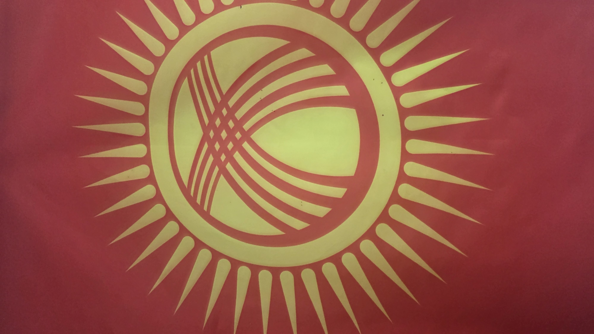 The image shows a close-up of the flag of Kyrgyzstan. The flag is red with a yellow sun in the center. The sun has 40 rays. Inside the sun is a yellow representation of a tunduk, which is the crown of a traditional Kyrgyz yurt. The tunduk is depicted with four wooden beams that support the roof of the yurt. The flag appears to be made of fabric and is displayed outdoors. The image does not provide any information about the location in the city of Tokmok City, Kyrgyzstan.