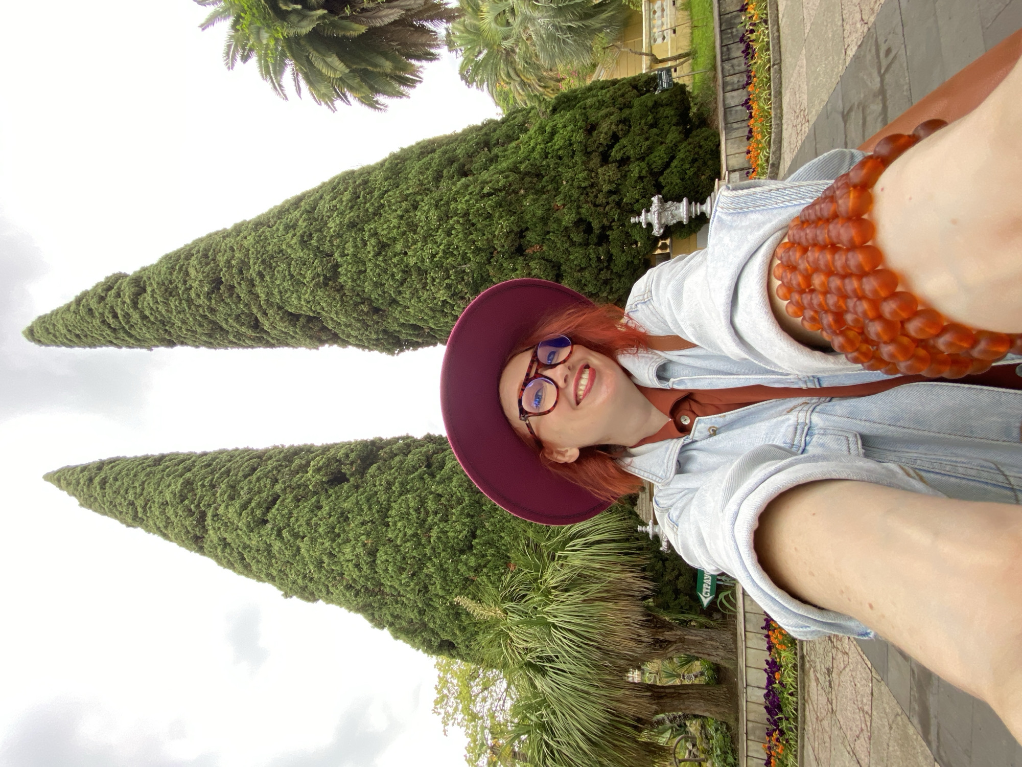 A selfie depicts a smiling red-haired woman with glasses and a wide-brimmed hat, holding her arm out toward the camera, displaying a large, amber-colored bead bracelet. She is wearing a denim jacket over a brown shirt. The background features two tall, cone-shaped trees. Also visible are palm trees, a fountain, a stone walkway, and flower beds with purple and orange flowers. A sign with Cyrillic text, possibly "СТРАУС," is partially visible. Based on the flora, the scene appears to be in a park or garden, likely in Sochi, Russia.