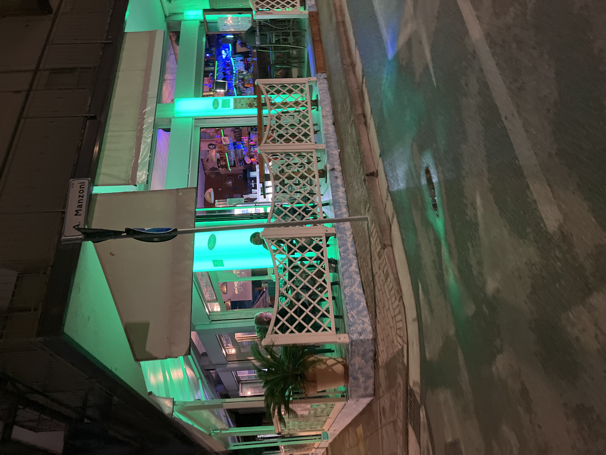 The image captures an evening scene on A. Manzoni street in Jesolo, Italy. A building, illuminated with green lights, houses a bar or restaurant with a seating area enclosed by white lattice fencing. Inside, multiple people are visible, some sitting and others possibly moving around. A potted palm plant sits on the patio. The street, wet and reflecting the green light, stretches into the distance. A street sign reading "A. Manzoni" is attached to a lamppost. The overall atmosphere is urban and vibrant due to the bright lights and activity within the building.