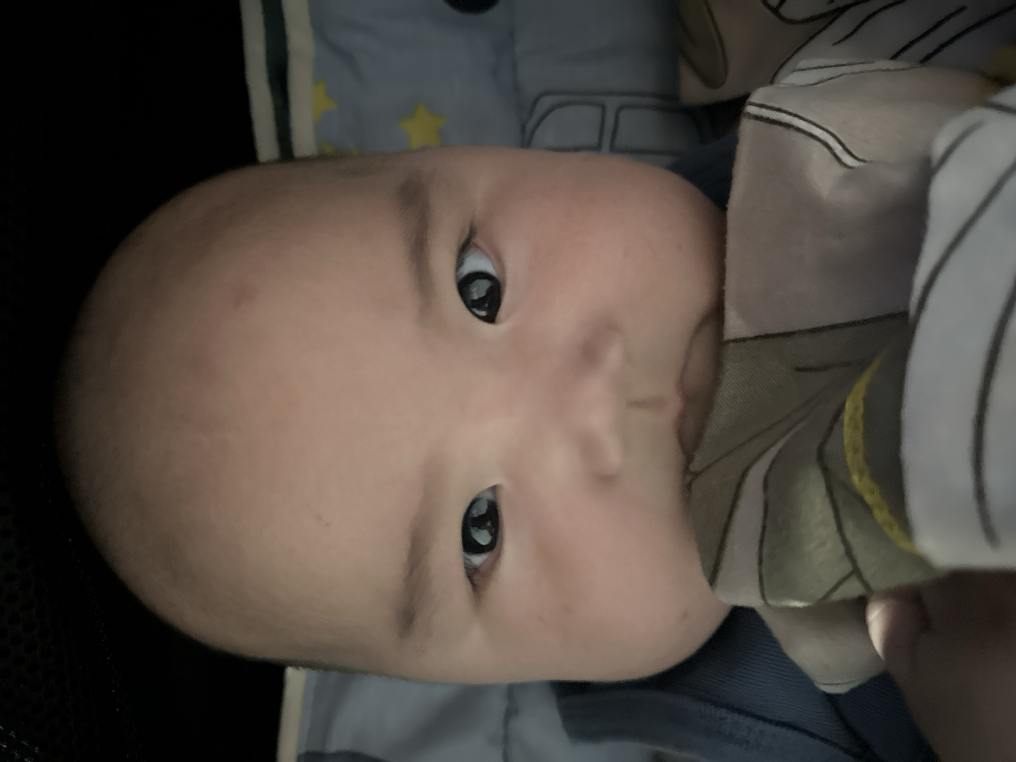 The image is a close-up, head-on, portrait-style photograph of a young baby's face. The baby is looking directly at the camera with wide, dark eyes. The baby's head is bald, and their cheeks are full and rounded. A portion of a patterned fabric is visible in the lower part of the frame, partially obscuring the baby's mouth. The background is dark and out of focus, with some hints of a patterned bedding or fabric to the right. There is no discernible location in the city of Тайгараев, Kyrgyzstan that can be identified from the image.