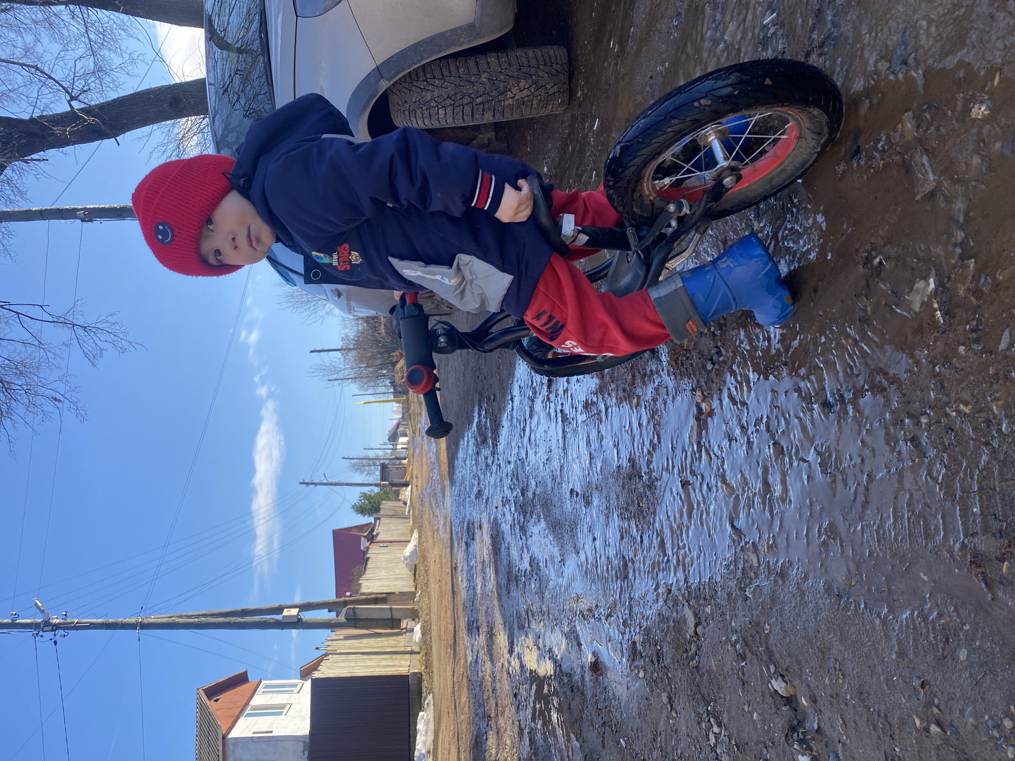 A young child in a red beanie, a blue jacket with patches, red pants, and blue rain boots is seated on a black balance bike on a muddy street. The street is flooded with water. The child is facing the camera. To the child's left is the lower portion of a silver-colored vehicle. In the background are wooden fences, buildings, and utility poles against a blue sky with a thin white cloud. The buildings and fences appear to be of the rural type. The location is identified as Ольховка, Russia.