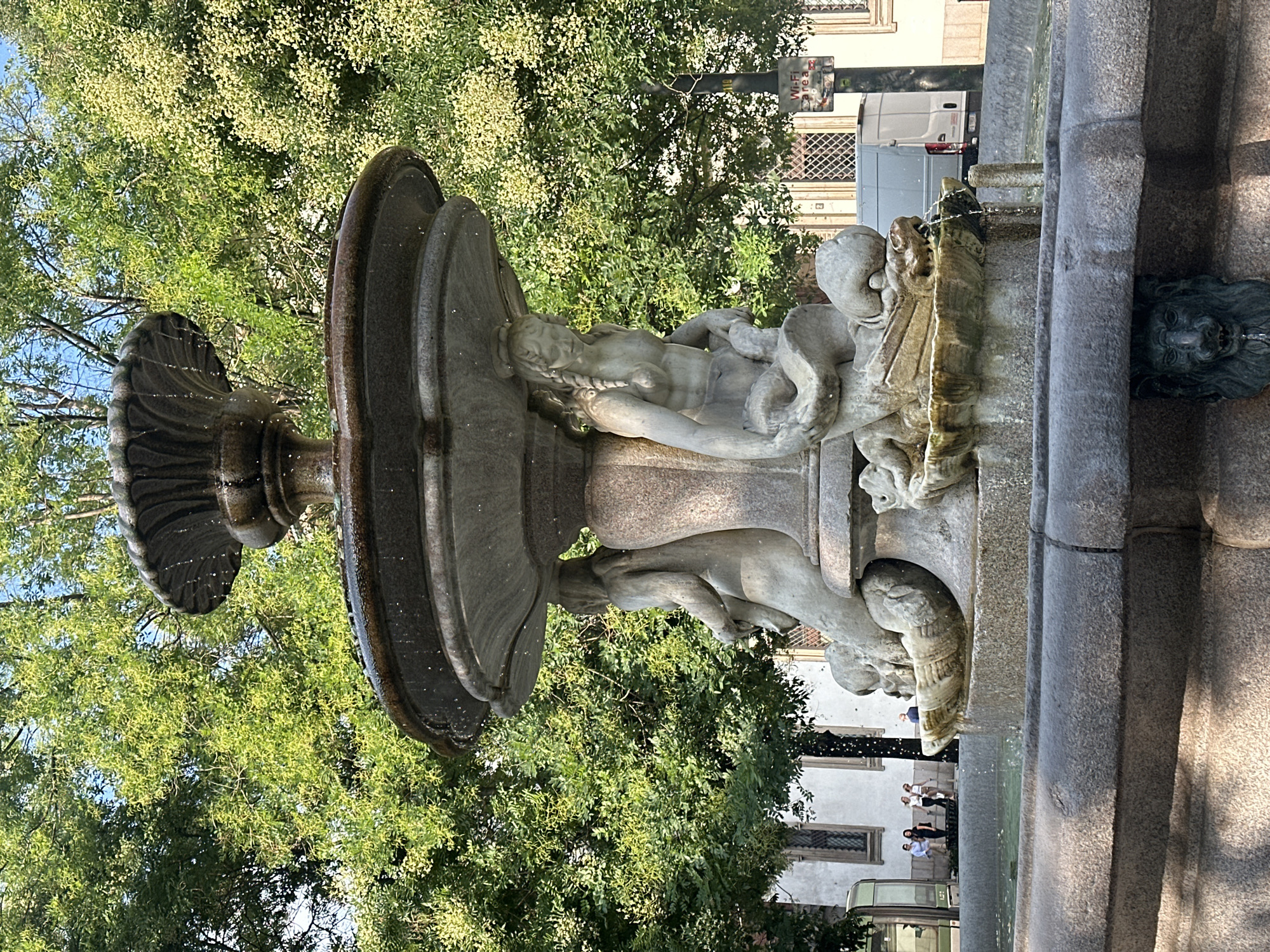 The image depicts a weathered stone fountain adorned with sculptures of mermaids and marine creatures. Water cascades from its tiered basins, pooling in the lower sections. In the background, lush green trees with dappled sunlight create a vibrant natural backdrop. To the left, a white building with arched windows is visible, and a few individuals are standing near it. Further back, a white van is parked. The fountain is situated in a public area, likely a square or park, given the presence of people and the architectural elements. The scene is set in Milan, Italy.