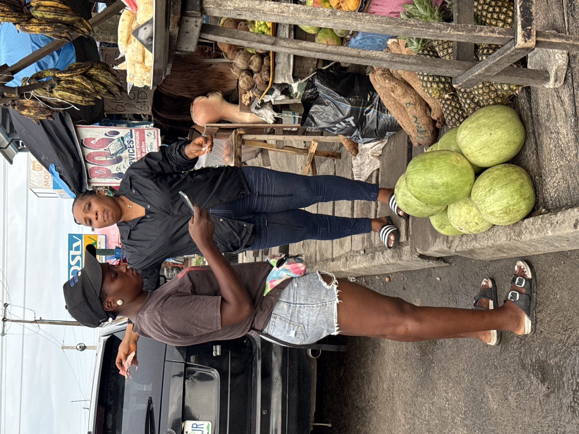 Two women are next to a black SUV in Ado, Nigeria. One woman wears a black shirt, dark jeans, and striped sandals. She holds a cell phone. The other wears a gray shirt, denim shorts, a black baseball cap, and black sandals. The SUV is parked on a street next to a roadside market stand. The market stand has plantains hanging, yams, watermelons, pineapples, and other items. A sign is also visible at the market. The sky is overcast.