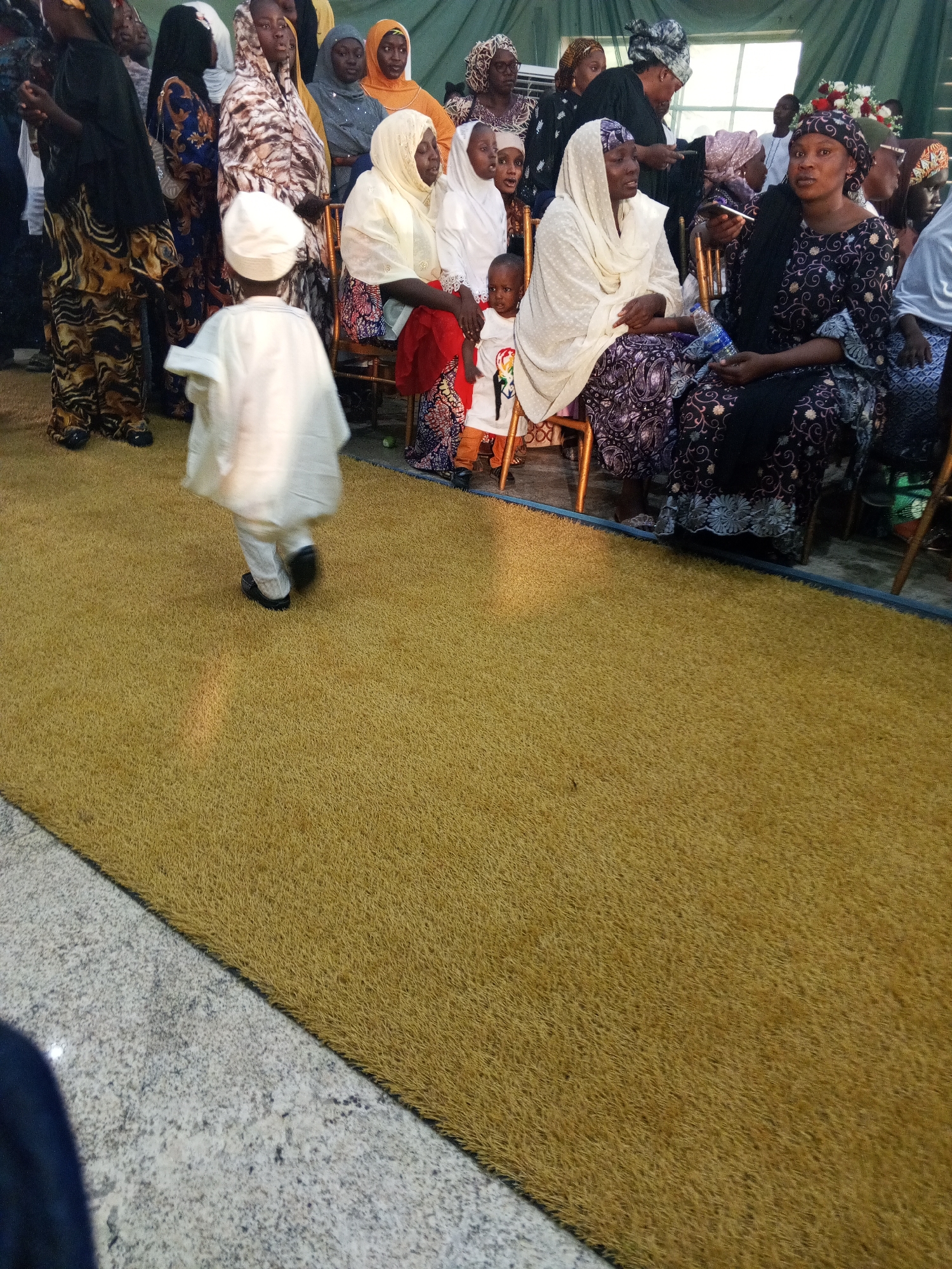 The image shows an indoor gathering with a group of people, mostly women and children, seated on chairs and standing. A young boy in a white outfit and cap is walking away from the camera on a yellow carpet. Several women are wearing hijabs and traditional clothing. Some are looking towards the camera, while others are engaged in conversation or looking elsewhere. The environment appears to be a hall or a room with green curtains in the background and a window. The lighting suggests it is daytime. The location is unknown.