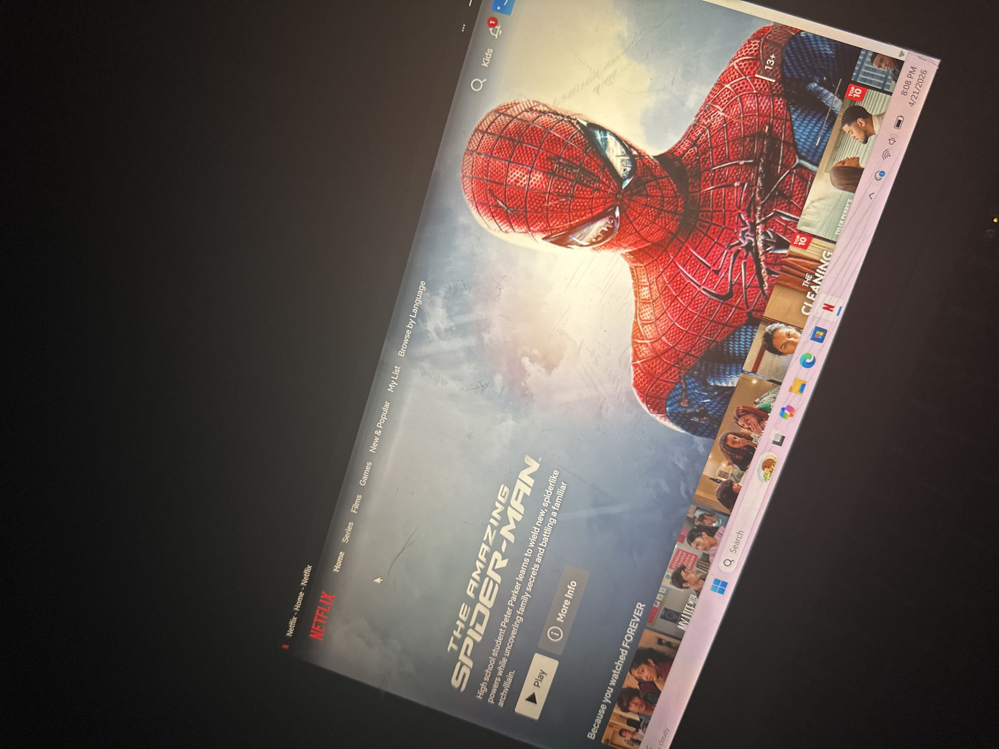 An image displays a computer monitor showing the Netflix streaming service, with "The Amazing Spider-Man" featured prominently. The screen's main subject is a large image of Spider-Man in his iconic red and blue suit. Digital text on the screen includes the movie title "THE AMAZING SPIDER-MAN," a synopsis ("High school student Peter Parker learns to wield new, spiderlike powers while uncovering family secrets and battling a familiar archvillain."), and "Play" and "More Info" buttons.
The Netflix interface shows navigation options such as "Home," "Series," "Films," "Games," "New & Popular," "My List," and "Browse by Language." A "Kids" profile icon is visible at the top. To the right, a sidebar displays various movie/show thumbnails, some marked "TOP 10" and a "13+" rating. Titles like "TYLER PERRY'S" and "THE CLEANING" are partially visible on suggested content.
The bottom right corner of the screen's operating system taskbar shows the time as "8:08 PM" and the date "4/21/2026," with a weather icon and the word "cloudy." Windows and Netflix application icons are visible on the taskbar.
The surrounding area of the screen is dark, suggesting an indoor setting. While the provided location context is Kirana, Nigeria, the digital content displayed on the screen is generic to the Netflix platform and Windows operating system, offering no specific visual cues unique to that geographical location. The activity depicted is the browsing or presentation of streaming entertainment content.