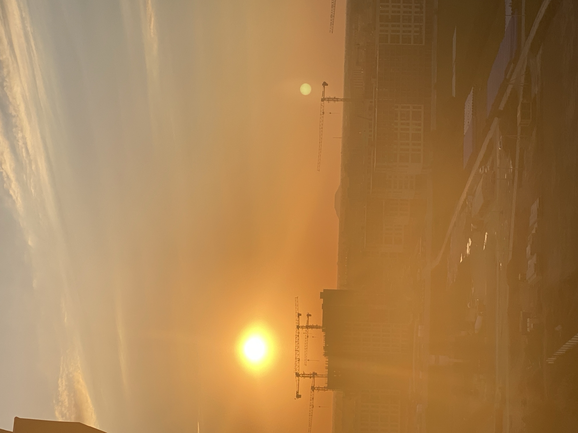 An aerial shot captures a city at sunset. The sky is a vibrant orange, with thin, wispy clouds scattered across it. The sun, a bright orb, dominates the upper left quadrant of the image, casting a warm glow over the scene. In the mid-ground, a cluster of construction cranes stands tall against the horizon, silhouetted by the setting sun. Beneath them, buildings are under construction, their skeletal structures partially visible through the haze. In the foreground, more buildings and what appears to be a construction site are visible, with roads and various structures scattered across the landscape. The overall impression is one of an urban environment in development, bathed in the dramatic light of a sunset.