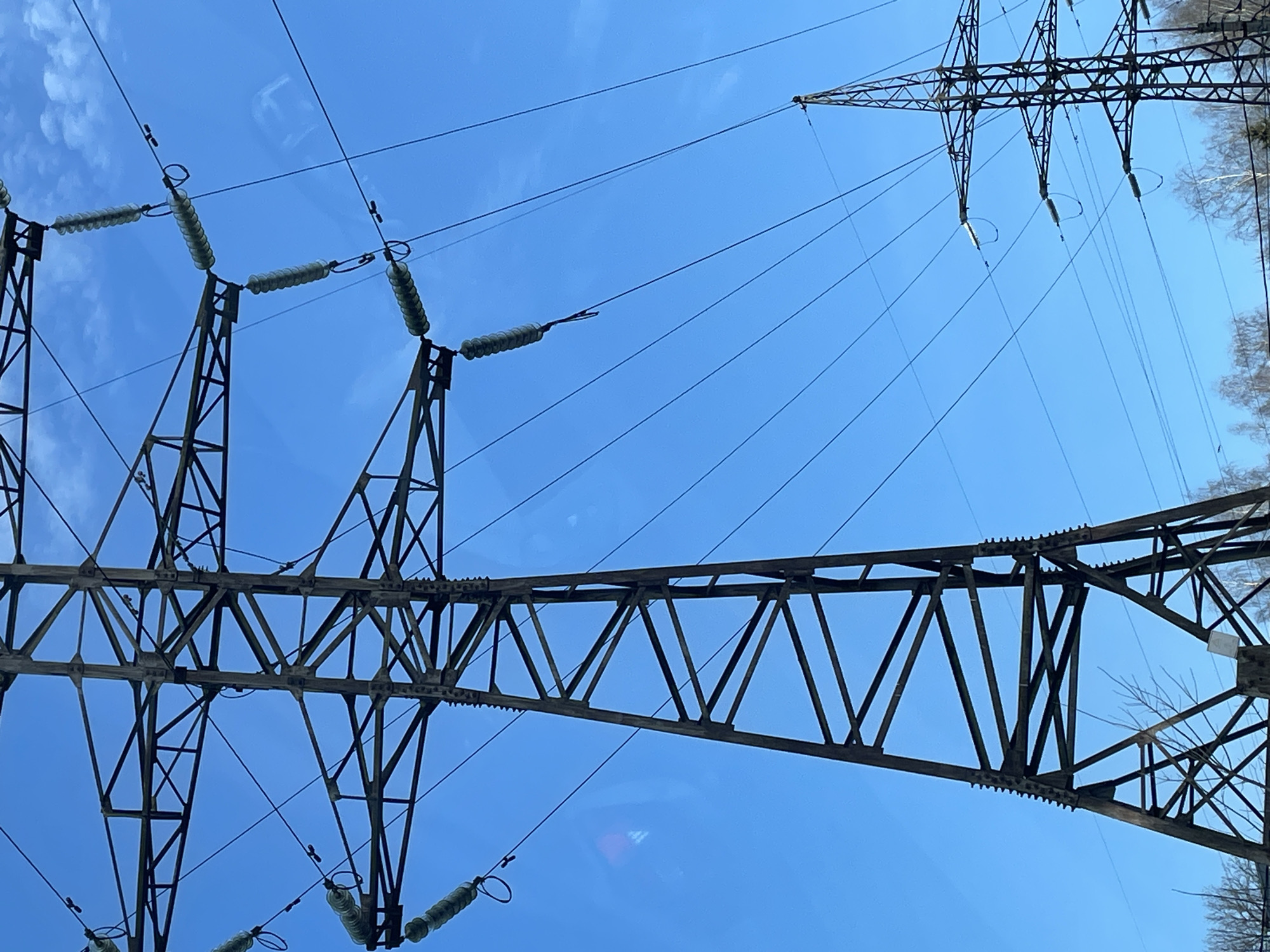 The image displays a low-angle view of two high-voltage electrical transmission towers against a predominantly clear blue sky in Izhevsk, Russia. The prominent tower, a large lattice-steel structure, occupies the left and central portion of the frame. It is constructed from dark, possibly weathered, metal and features multiple horizontal cross-arms. Attached to these arms are multiple strings of translucent disc insulators, from which at least six parallel power lines extend horizontally and diagonally across the frame. A second, smaller lattice-steel transmission tower is visible in the distant lower right, also supporting power lines. The sky is a vibrant blue with sparse, wispy white cloud formations in the upper right quadrant. The bare branches of deciduous trees and the tops of some evergreen trees are visible along the lower edge of the frame. A small, white rectangular sign is affixed to a diagonal strut near the base of the larger pylon. No individuals, vehicles, or specific human activities are present.