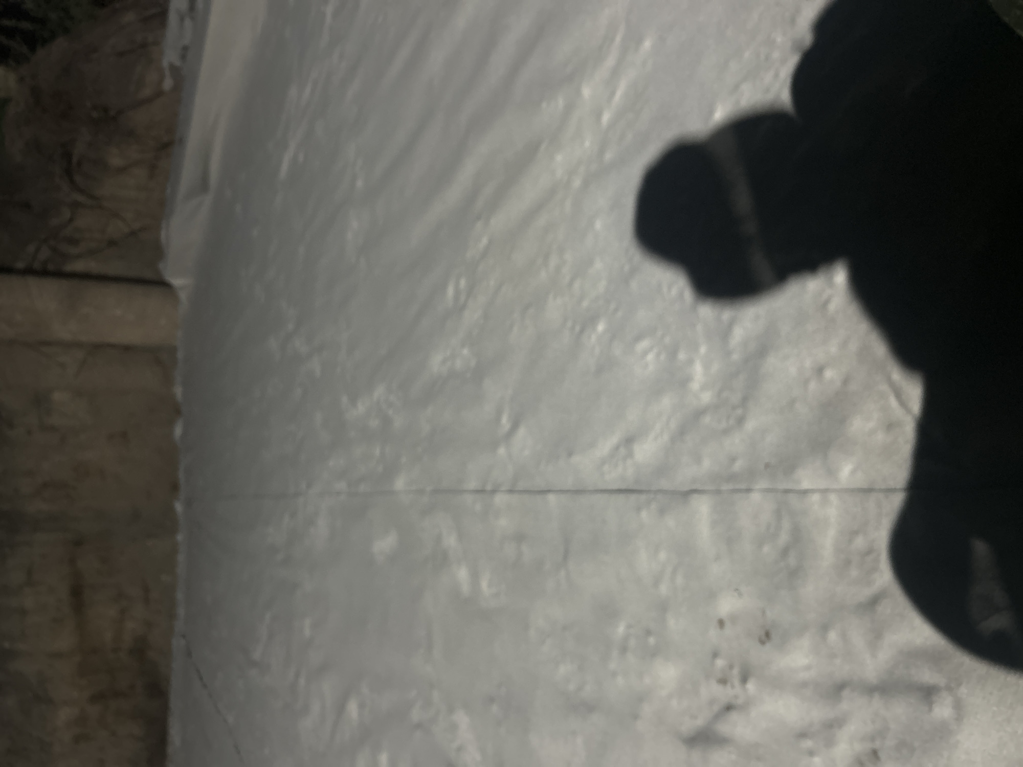 The image depicts a shadowy silhouette of a person wearing a hat and a mask, seen from behind, against a backdrop of a snow-covered slope. The scene is likely outdoors, possibly in a park or a recreational area within the city of Krasnodar, Russia, given the context of snow. The ground is covered in a thick layer of snow, with visible tracks or lines suggesting movement across the surface. The background is dimly lit, with what appears to be a stone wall or embankment. The overall atmosphere is nocturnal or dusk, with limited natural light.