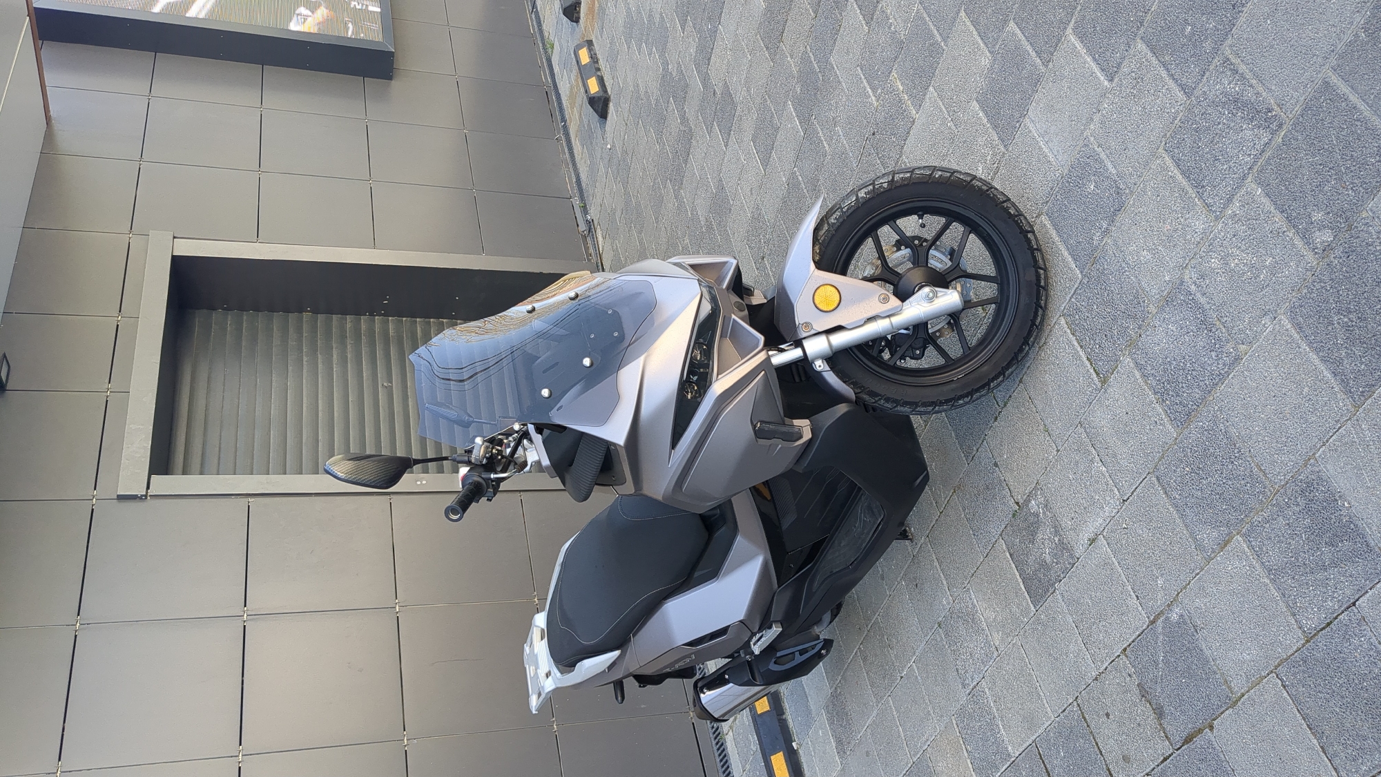 A modern, grey-silver scooter is prominently parked on a paved surface next to a contemporary building. The scooter faces slightly left and forward, showcasing its sleek design. It features a dark tinted windshield, integrated LED-style headlights, a black two-tone seat, and one visible rearview mirror with a carbon fiber-like pattern on the left handlebar. The front wheel has a black rim and silver-colored suspension components, with a round yellow turn signal positioned above it.
The ground is covered with grey interlocking pavers arranged in a diagonal pattern. To the left, a dark grey building with large square wall tiles serves as the background. A recessed section within the building features a corrugated or ribbed interior. Above this, a large rectangular sign or display reflects the surroundings, but no discernible text is visible. Black road barriers with yellow reflective strips are present further in the background. The scene is illuminated by bright daylight, suggesting clear and sunny weather. No people or specific activities are depicted. The setting is an urban or commercial area in Gelendzhik, Russia.