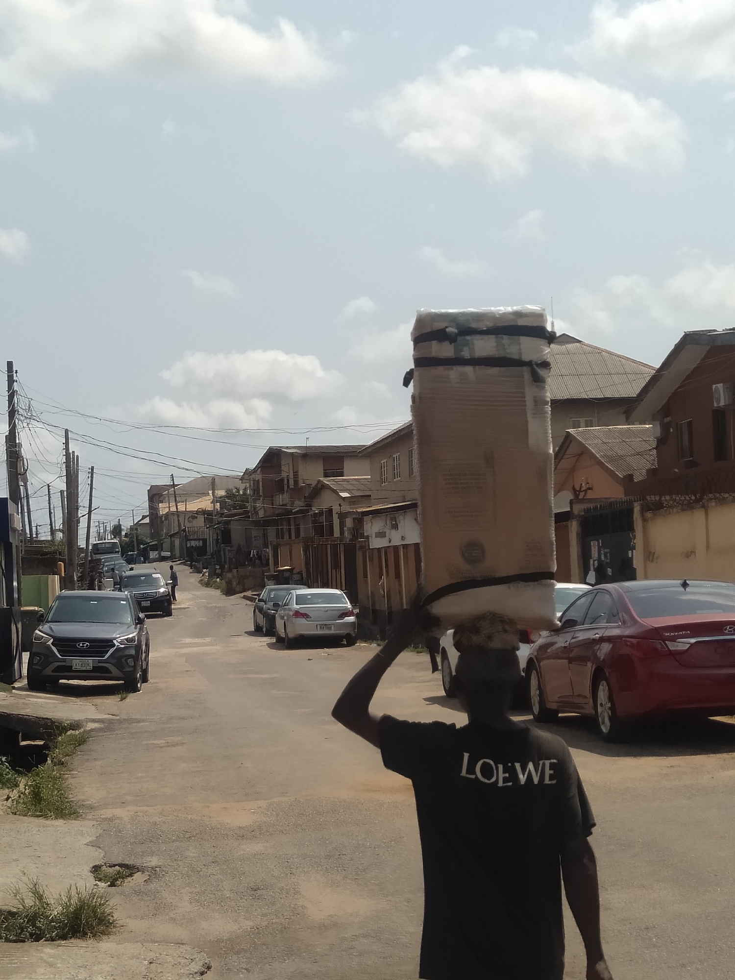 In Shangisha, Nigeria, a man wearing a black T-shirt with "LOEWE" printed on the back is carrying a large cardboard box wrapped with plastic and tape on his head. He walks down a street lined with parked cars, including a gray Hyundai, a silver sedan, and a red Sonata. The street also has drainage systems, telephone poles, and a mix of residential buildings. The sky is overcast with scattered clouds.