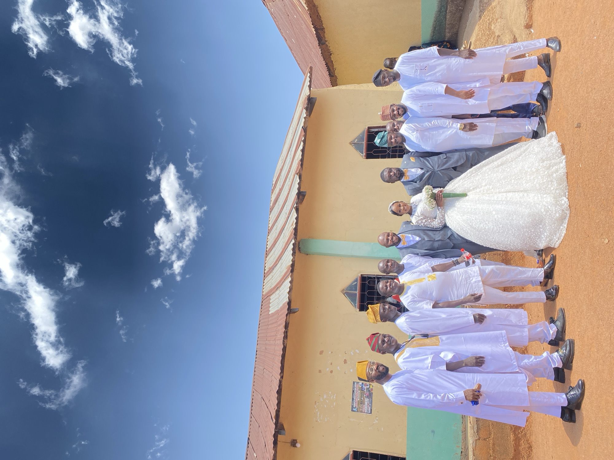 The image shows a wedding group posing in front of a one-story building in Vom, Nigeria. The bride is wearing a white lace wedding dress, holding a bouquet, and is flanked by men in grey suits with yellow bow ties. The other men are dressed in white traditional outfits with colored hats. The building has a rusty, corrugated metal roof, yellow walls, and green trim. The sky is blue with scattered white clouds.