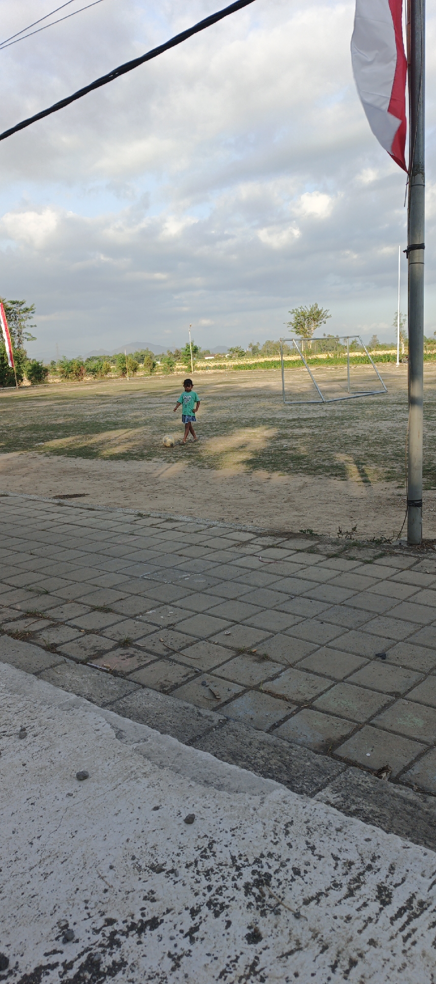 In Kasiyan Timur, Indonesia, a young child, likely a boy, is observed on a dirt and grass field, wearing a teal t-shirt and blue shorts. He is standing next to a deflated soccer ball, suggesting he was engaged in play or has just finished. A small, metal soccer goal is visible in the background, indicating the area is likely a sports field or park. The sky above is partly cloudy, with a flag pole topped by a partially visible Indonesian flag to the right of the frame, and another flag to the left. The foreground is dominated by a paved walkway made of square concrete tiles. The overall environment appears to be an outdoor public space, possibly a community field or schoolyard, under a cloudy sky.