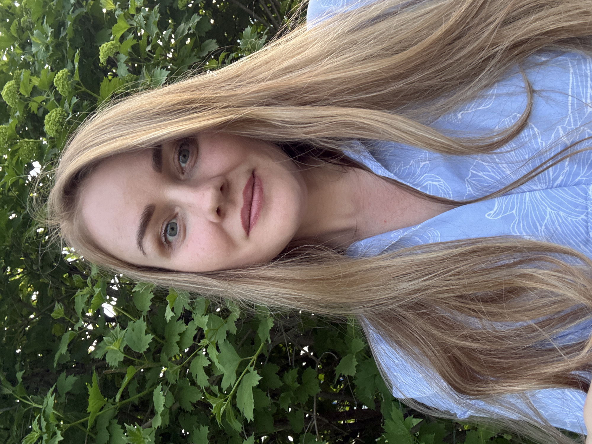 A young woman with long blonde hair and light eyes is the focal point of the image. She is wearing a light blue shirt with a floral pattern. Her face is visible, and she appears to be smiling slightly. The background consists of green foliage, potentially a tree or shrubs. The image appears to be taken in an outdoor setting. There is no verifiable information to determine if this image was taken in Chaikovsky, Russia.