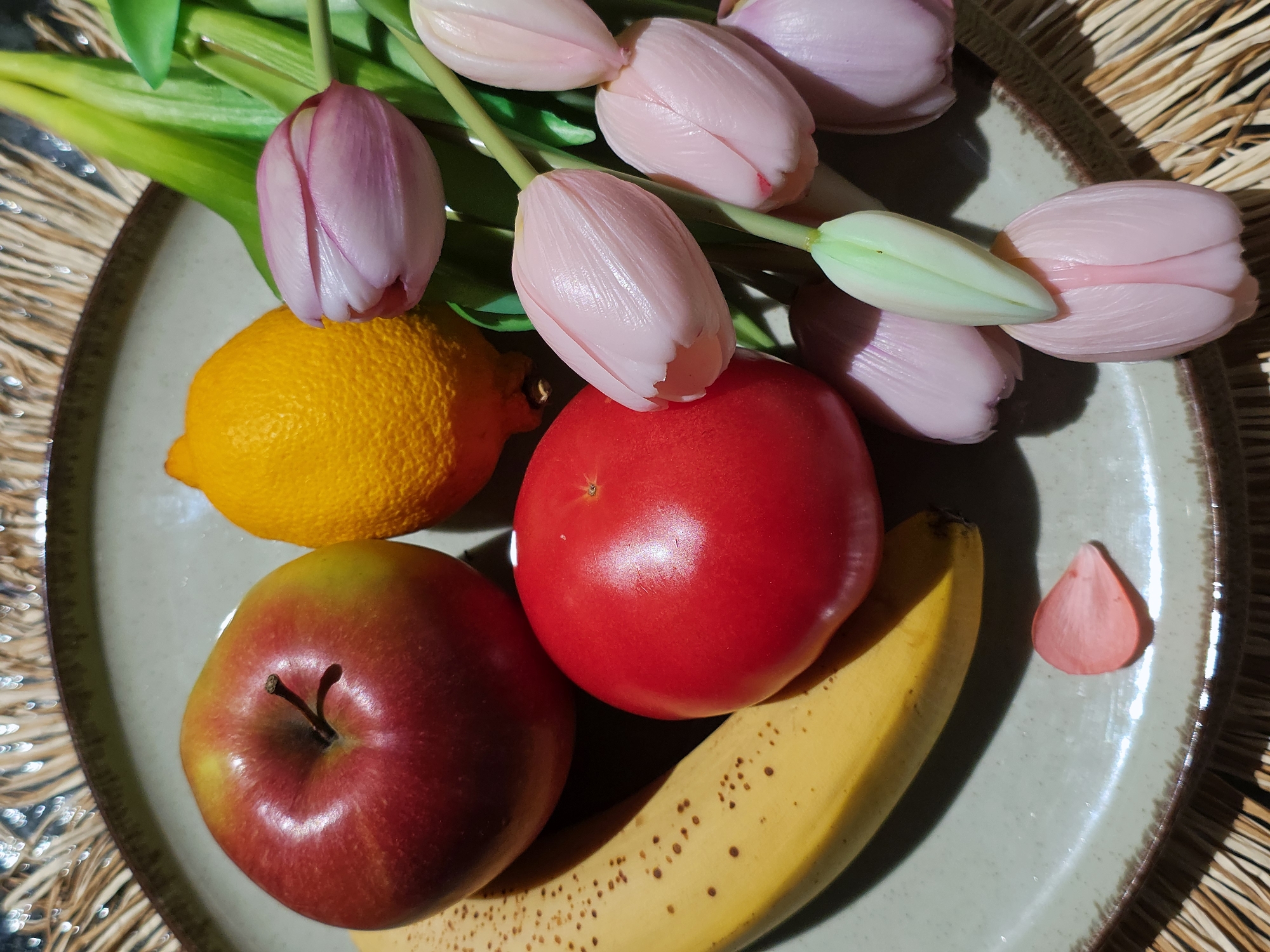 A still-life photograph shows a plate filled with fruit and flowers. The plate, which has a neutral beige tone with a dark rim, holds a yellow lemon, a red apple, a red tomato, and a yellow banana. There are also several pink tulips with green stems. A single petal, possibly from one of the tulips, sits on the plate. The plate is positioned on a woven surface. The location is unknown.