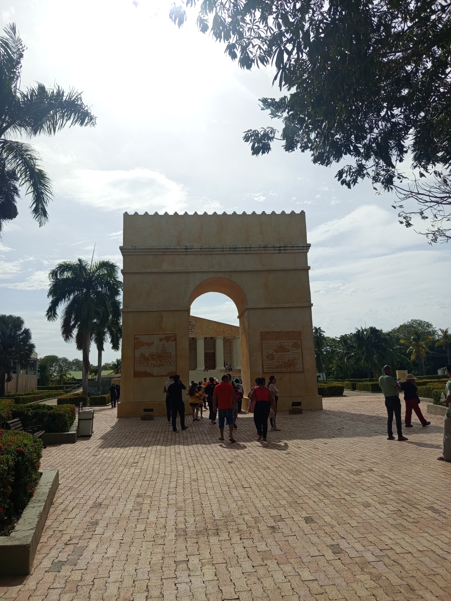 In Coveñas, Colombia, a broad, sun-drenched stone pathway leads towards a grand, beige archway. The archway features a crenelated top and intricate bas-relief panels on its facade, depicting historical scenes. Beyond the arch, a building with classical columns is visible. Palm trees and lush green landscaping surround the area, with a bright, partly cloudy sky overhead. Several individuals are present, walking along the path and gathering near the archway. The scene suggests a tourist destination or historical site.