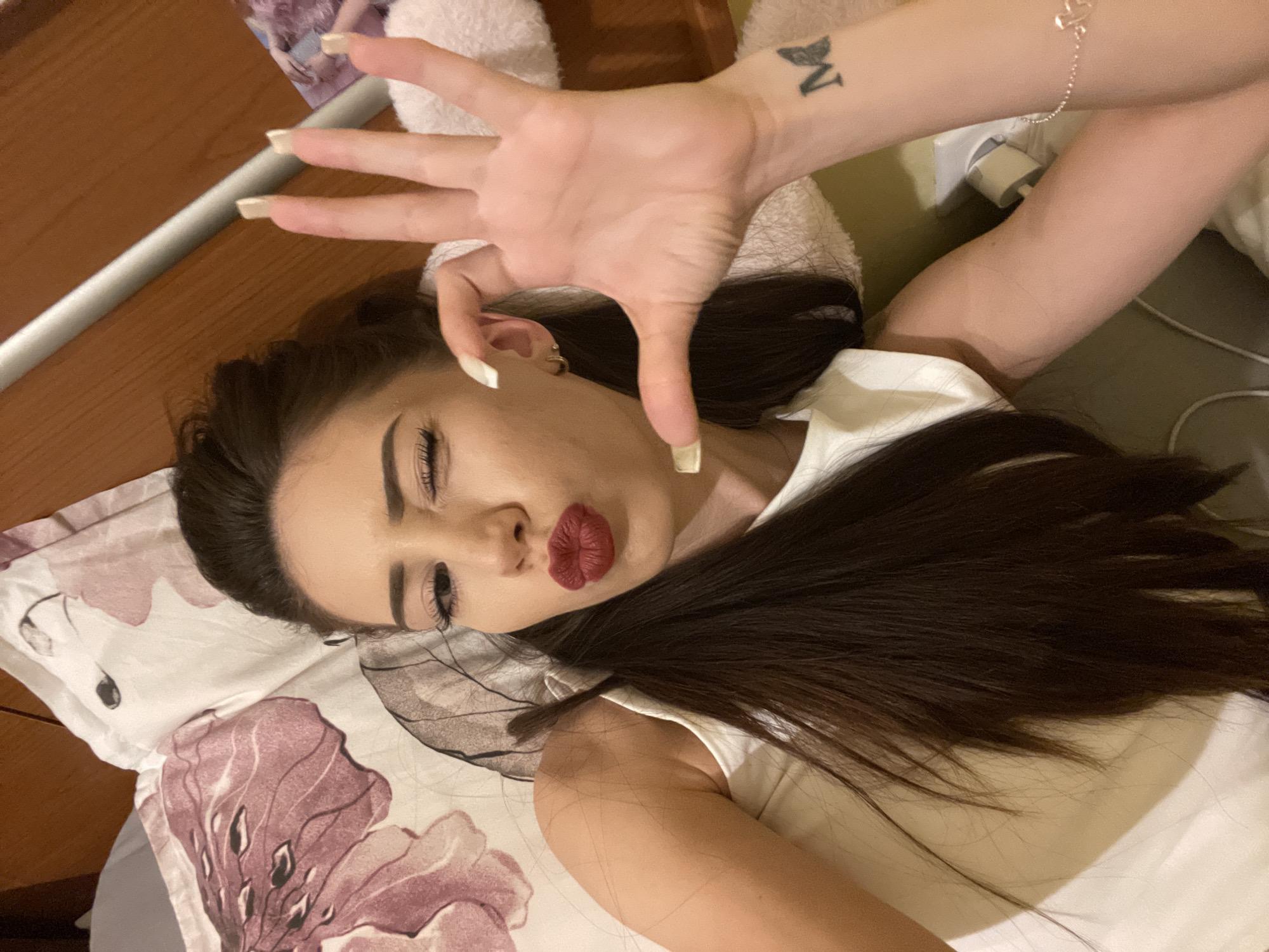 A woman with dark hair and dark lipstick winks at the camera while lying on a bed. Her right hand is held up, obscuring part of her face. She wears a white top and a bracelet. A tattoo of the letter "M" is visible on her left forearm. The background shows a floral-patterned pillow and a wooden headboard.