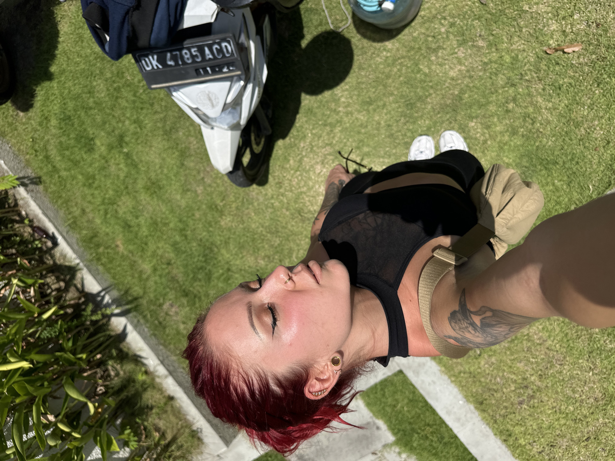 The photograph is taken from an elevated perspective, looking down on a scene set in Dalung, Indonesia. A woman with short, dyed red hair, multiple ear piercings, and tattoos on her arms is the primary subject. She is wearing a black crop top and black leggings, with white sneakers visible near her feet. A light brown backpack or bag is slung over her shoulder.
To the left of the woman is a white scooter parked on a patch of green grass. The scooter has a black license plate with the inscription "DK 4785 ACD". A dark blue bag is strapped to the front of the scooter. A clear helmet rests on the grass near the scooter.
The environment consists of green grass and foliage, suggesting an outdoor setting. A concrete walkway is visible on the left edge of the frame, indicating the presence of a building or structure nearby. The scene is brightly lit, suggesting it is daytime.