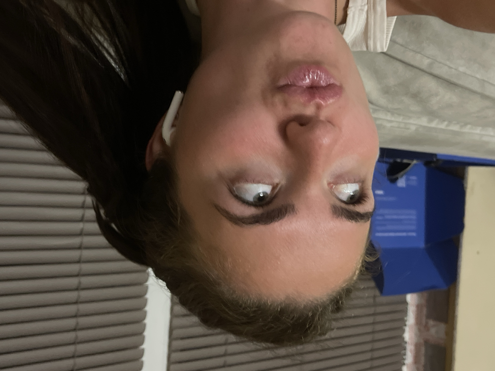 A young Caucasian woman is pictured from the chest up, looking upwards and to her left. She has dark brown hair pulled back in a ponytail, fair skin, and is wearing a white ribbed tank top. She has on white wireless earbuds in her right ear. Her lips are pursed. In the background, to the left, a blue box is visible, and behind her is a wall with horizontal blinds. The setting appears to be indoors, possibly a residential interior. The location in the city of Мосметаллоконструкция Онт, Russia, is not discernible from the image.