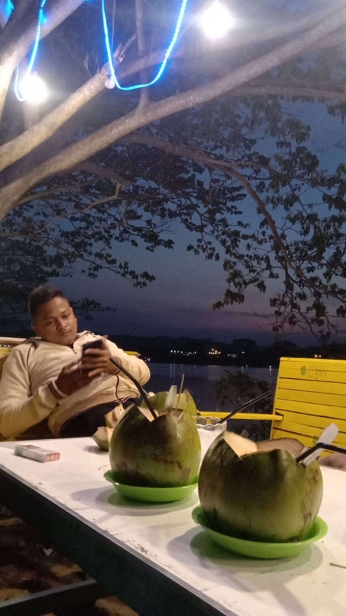 The image depicts a man relaxing outdoors at dusk or early night in what appears to be Lhokseumawe, Indonesia. He is seated at a white table, dressed in a light-colored, long-sleeved zippered top, and is engrossed in his smartphone, which he holds with both hands.
On the table in front of him are two green young coconuts, each opened at the top and served with a straw and small spoon, resting on bright green plastic saucers. A red and white cigarette pack, identifiable as Marlboro, lies on the table to the left of the coconuts. Partially visible to the right is a yellow slatted chair.
The setting overlooks a calm body of water, likely a lake or river, with faint lights from the distant shore reflecting on its surface. The sky is a deep twilight blue, subtly transitioning to shades of purple and pink near the horizon. Large tree branches, with dark bark and sparse leaves, extend over the scene, adorned with bright white and electric blue string lights, illuminating the area. The overall atmosphere is peaceful and relaxed.