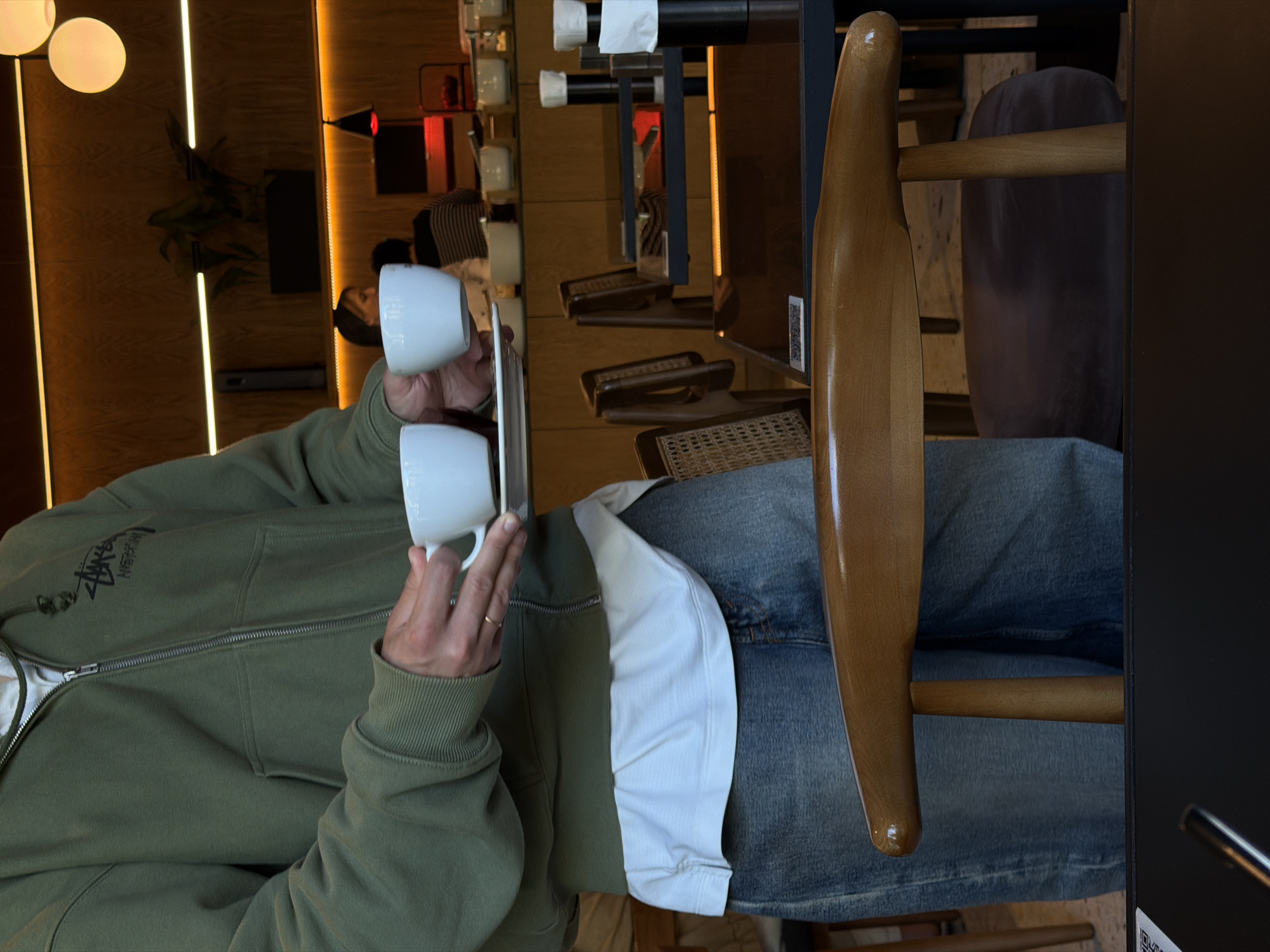 A person wearing an olive green zip-up hoodie and blue jeans sits in a chair in a café in Tbilisi, Georgia, holding two white coffee cups. The café features wooden paneling, pendant lights, and a row of small dining tables with cushioned chairs along the wall. There are also what appear to be napkin holders mounted on the wall.